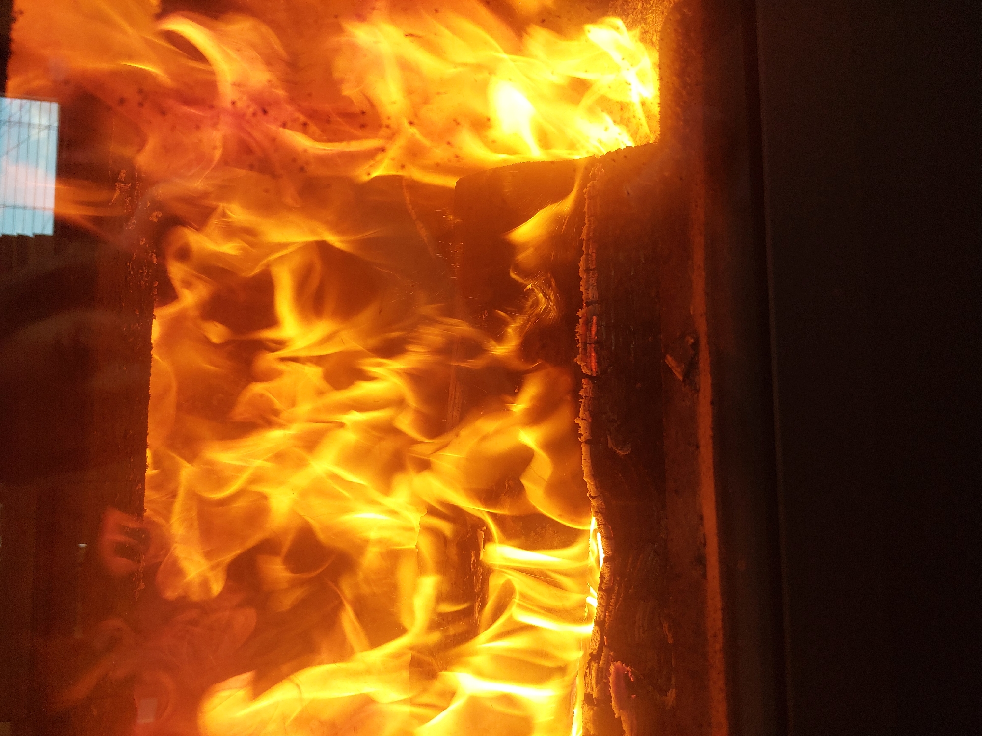 The image shows a fire burning in a wood-burning stove. The fire is intense and fills the frame, with bright yellow and orange flames. Pieces of wood are visible within the fire, and the stove's metal frame surrounds the flames. Reflected light is visible on the glass of the stove door. There is no visible reference to the location of Nedelino, Bulgaria.