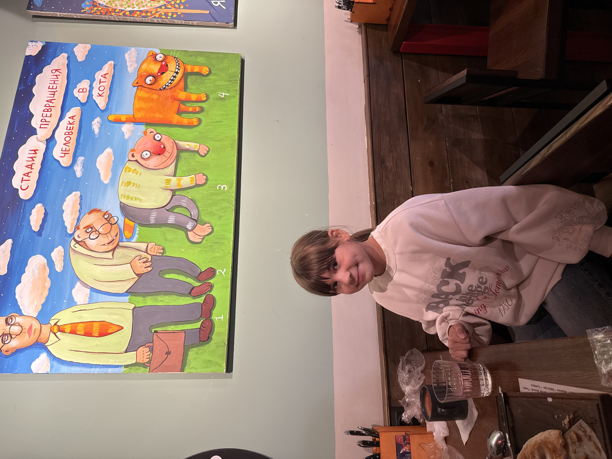 The photograph captures a young girl seated at a wooden table in what appears to be a restaurant or cafe in Заволжье, Russia. The girl, with light brown hair, is wearing a light-colored sweatshirt with some visible text "1980" and parts of "BICK" and "Tomorrus". She is looking towards the camera with a gentle smile, appearing content.
On the dark wooden table in front of her, there's a clear glass filled with liquid, a dark mug, some plastic wrap, cutlery, and a piece of paper or menu with Russian city names like "Ростов Великий" and "Чебоксари". The background features a light green wall adorned with a large, colorful painting.
The painting illustrates a whimsical concept titled "СТАДИИ ПРЕВРАЩЕНИЯ ЧЕЛОВЕКА В КОТА" (Stages of Human Transformation into a Cat), with numbers 1 through 4 marking the stages. The first stage shows a man in a business shirt and tie holding a briefcase. The subsequent stages depict his gradual transformation into a more relaxed, cat-like human, culminating in an orange, grinning cat. The overall setting suggests a casual, inviting atmosphere, likely during an evening meal or break.