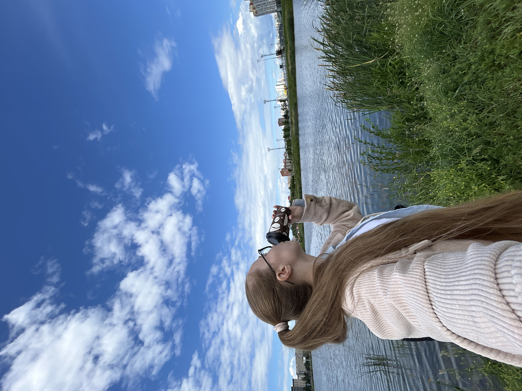 A young woman with long blonde hair in a ponytail, wearing glasses and a light pink corduroy jacket over a blue shirt, is standing by a body of water and drinking from a disposable coffee cup. The scene is outdoors with a bright blue sky dotted with white clouds. The water is calm with ripples, and lush green grass and reeds line the bank. In the background, across the water, there are buildings and some sports facilities with lights, indicative of an urban park or recreational area. The overall impression is a leisurely moment in an outdoor setting within a city. Based on the urban landscape, this could be a location like the park near the Murino residential complex, which features a similar body of water and modern buildings.