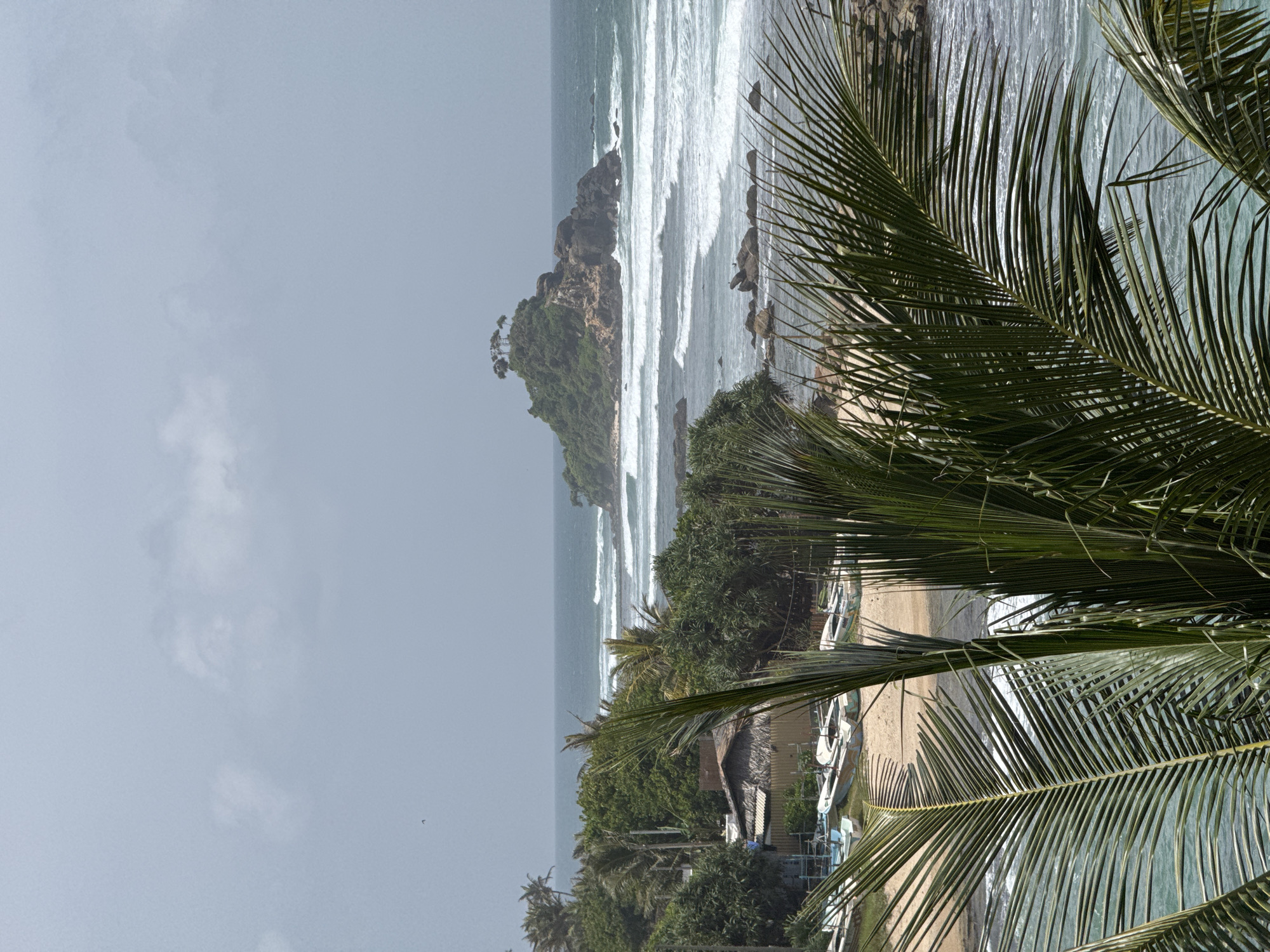 The image displays a coastal scene in Goviyapana, Sri Lanka, with lush greenery and the ocean. In the foreground, several palm fronds partially obscure the view, suggesting a tropical setting. Beyond the fronds, a sandy beach is visible, lined with several small fishing boats. A cluster of simple structures with thatched roofs are situated on the edge of the beach, indicating a local fishing village. The ocean exhibits moderate waves, with a prominent rocky outcrop or small island emerging from the water in the mid-ground. This island is topped with trees, adding to the natural beauty of the landscape. The sky is overcast with a light haze.