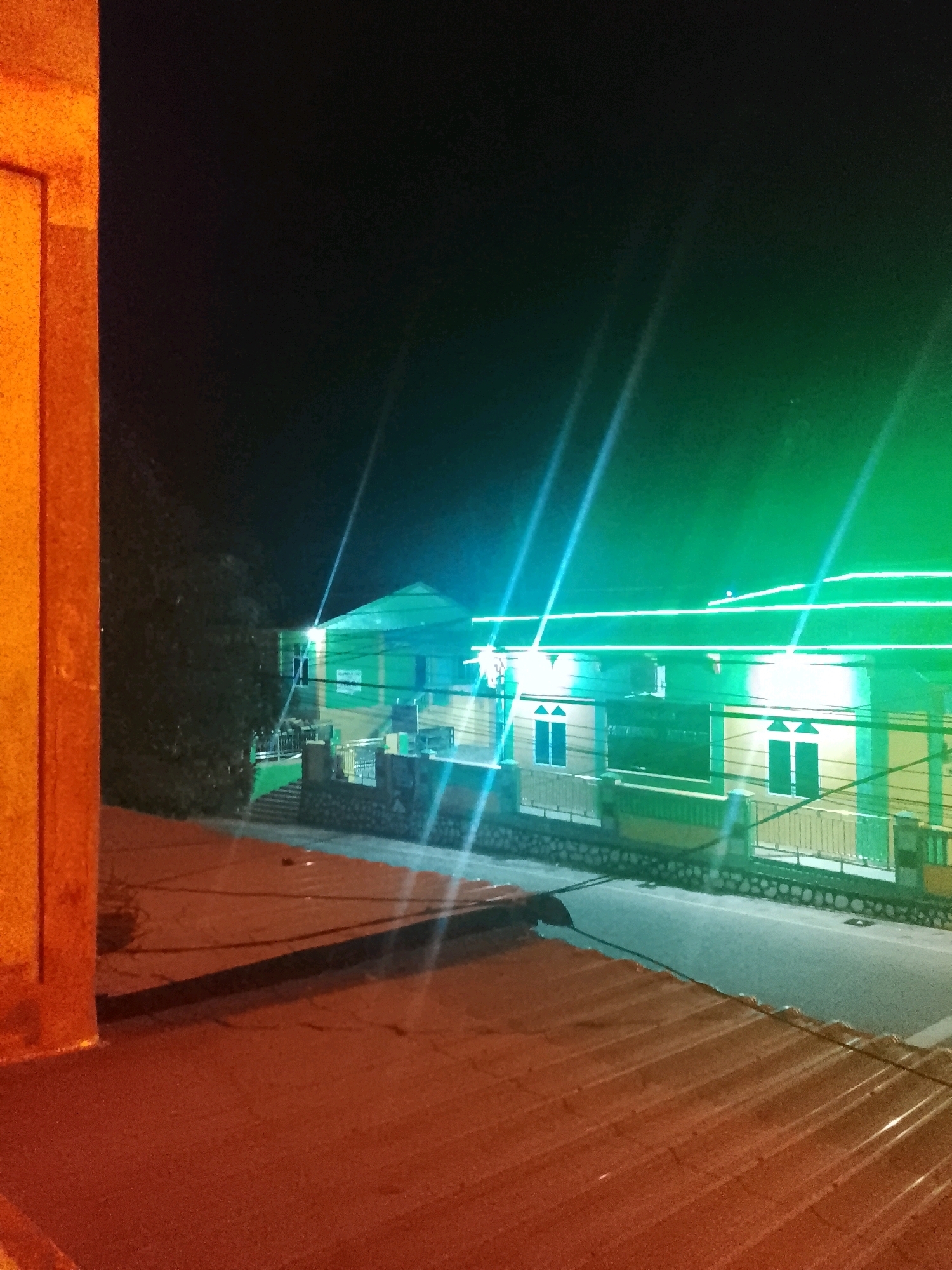 The image captures a nighttime view of a street in Tanjung Balai Karimun, Indonesia. The scene is illuminated by vibrant green and blue neon lights, which streak across the frame and highlight the architecture of the buildings. In the foreground, a reddish-brown corrugated metal roof is visible, suggesting a rooftop perspective.
The main subject is a multi-story building, likely a commercial establishment or residence, adorned with horizontal neon lighting along its facade. The building appears to be painted in a yellow and green color scheme. There are visible windows with dark shutters or panes. A stone wall and a metal fence line the front of the building. A paved street runs in front of the building, with some shadowy areas indicating the presence of parked vehicles or the absence of light. Trees are visible to the left of the building, partially obscuring it. The overall impression is one of a well-lit urban environment at night, with the neon lights creating a dynamic and somewhat artificial atmosphere.