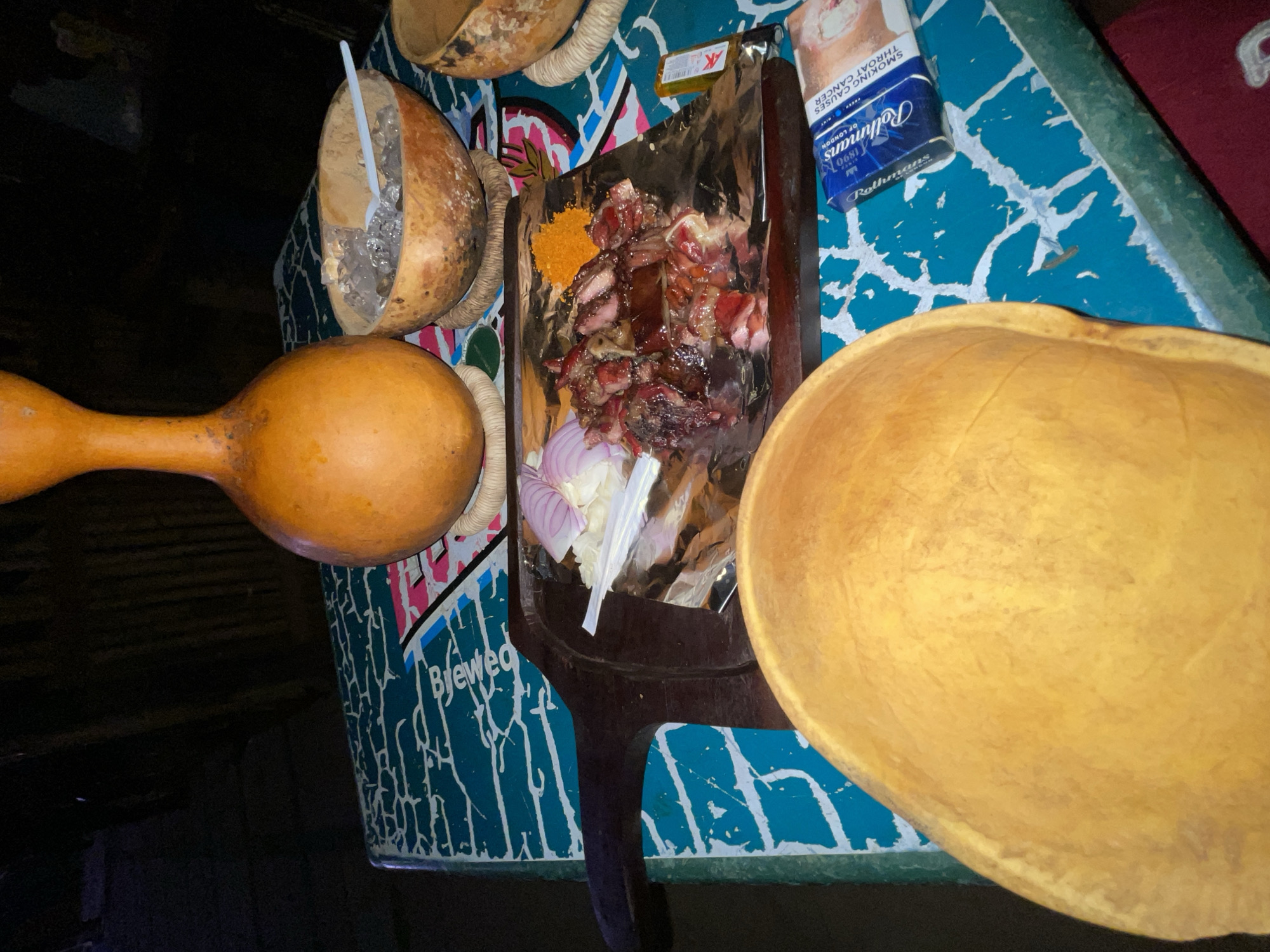 The image shows a table arrangement, likely in Abuja, Nigeria, featuring local cuisine and beverages. A platter of grilled meat (possibly suya) is displayed on aluminum foil, accompanied by sliced onions and a spice mix. A pack of Rothmans cigarettes is nearby. Traditional calabash bowls are present, one containing a plastic spoon and possibly ice. The table has a blue and white patterned surface, potentially indicating a local bar or eatery. A distinctive "Brewed" design is visible on the table surface, possibly referencing a local beer brand. The overall setting suggests a casual, local dining experience in Abuja.