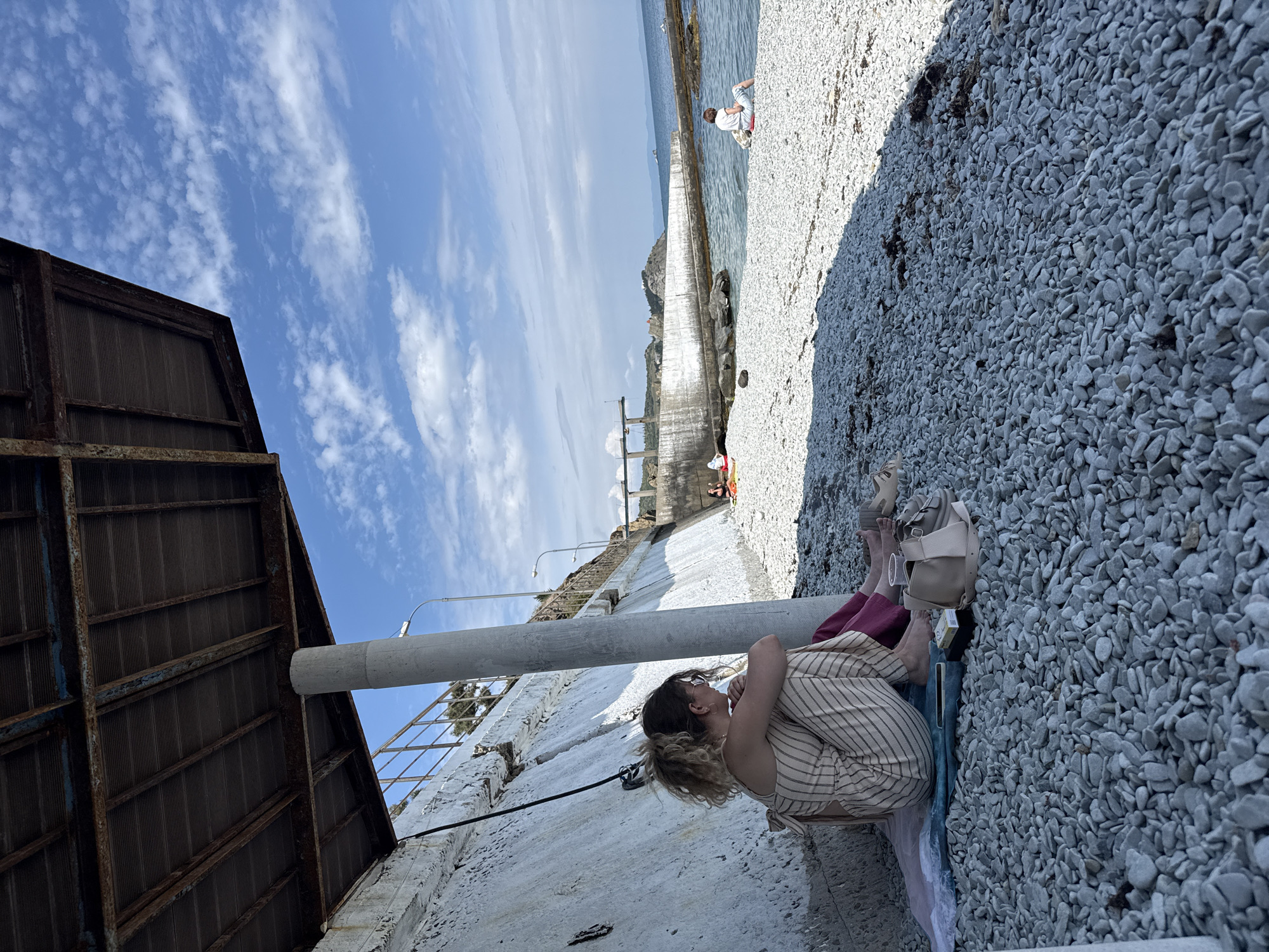 The image shows a woman sitting on a stony beach. The woman is wearing a striped dress and is sitting with her knees drawn up to her chest. A light-colored handbag and shoes are beside her. In the background, a concrete structure leads out to the sea, where a person is standing. The sky is blue with scattered clouds. The scene appears to be at a waterfront location.