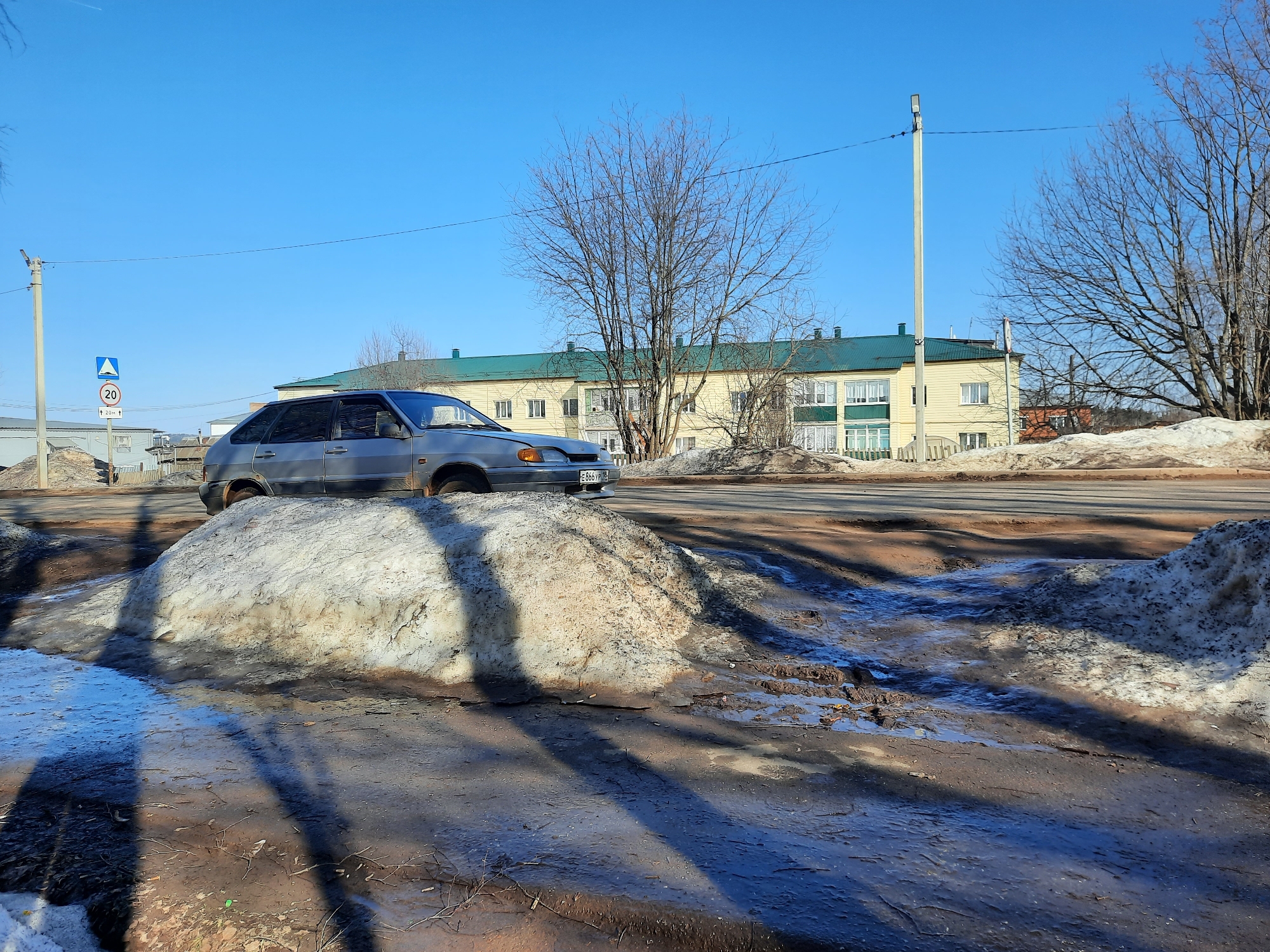 A silver car is positioned on a mound of melting snow along the side of a road. In the background, there is a yellow two-story building with a green roof. A speed limit sign is visible to the left of the car. Utility poles line the road. The scene takes place in Krasnogorskoye, Russia. The weather appears to be sunny. Bare trees are visible in the background. The ground is muddy with melting snow and puddles.