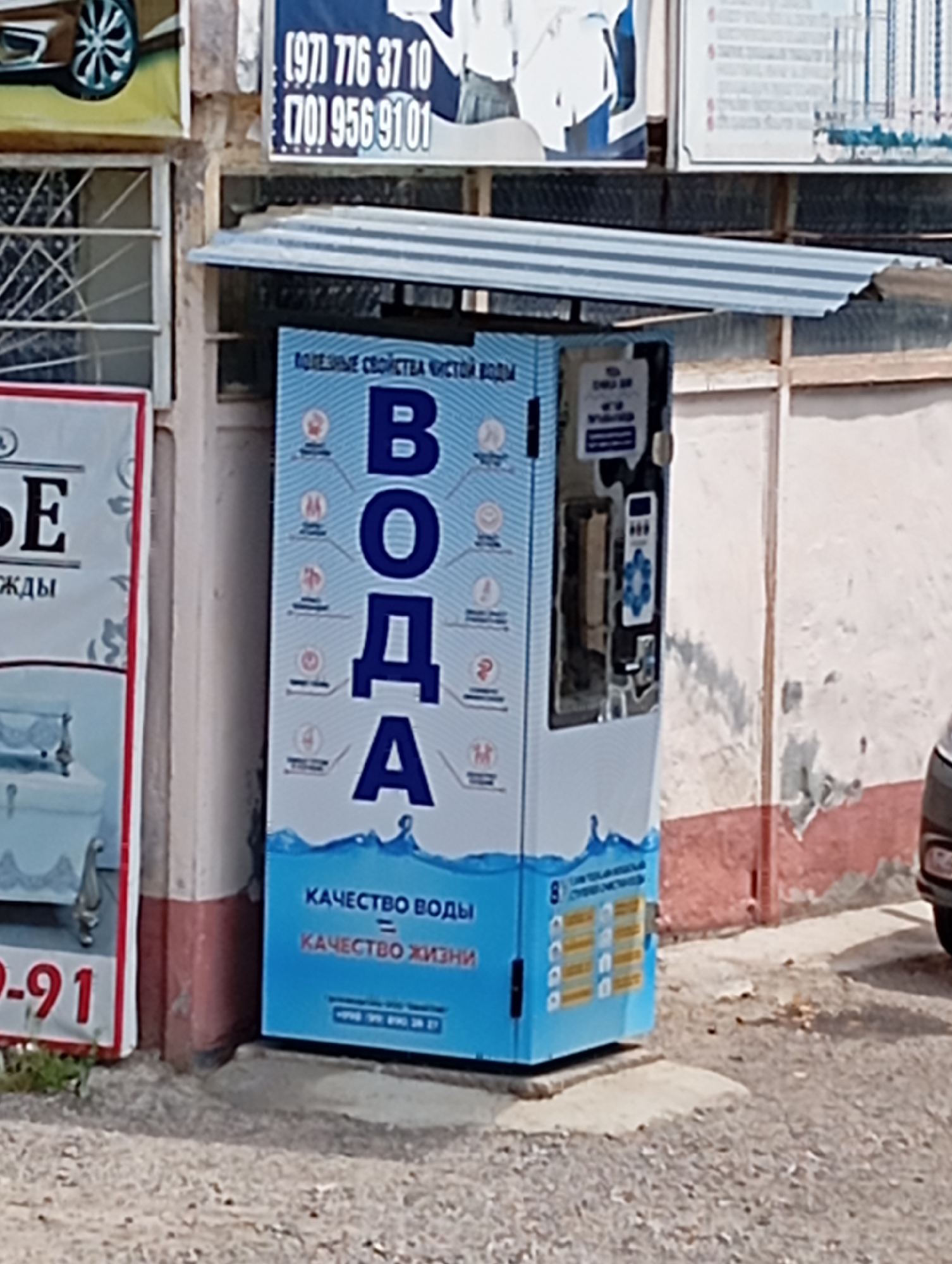 The image captures a water vending machine in Yukariyuz, Uzbekistan. The machine, colored blue and white, features the word "ВОДА" ("water" in Russian) prominently displayed, along with health-related icons and the slogan "КАЧЕСТВО ВОДЫ = КАЧЕСТВО ЖИЗНИ" ("water quality = quality of life"). It is situated next to a building with advertisements above and on its left side. The vending machine is protected by a corrugated metal roof. A car's headlight is partially visible on the right side of the image. The street in front of the machine is unpaved and dusty.