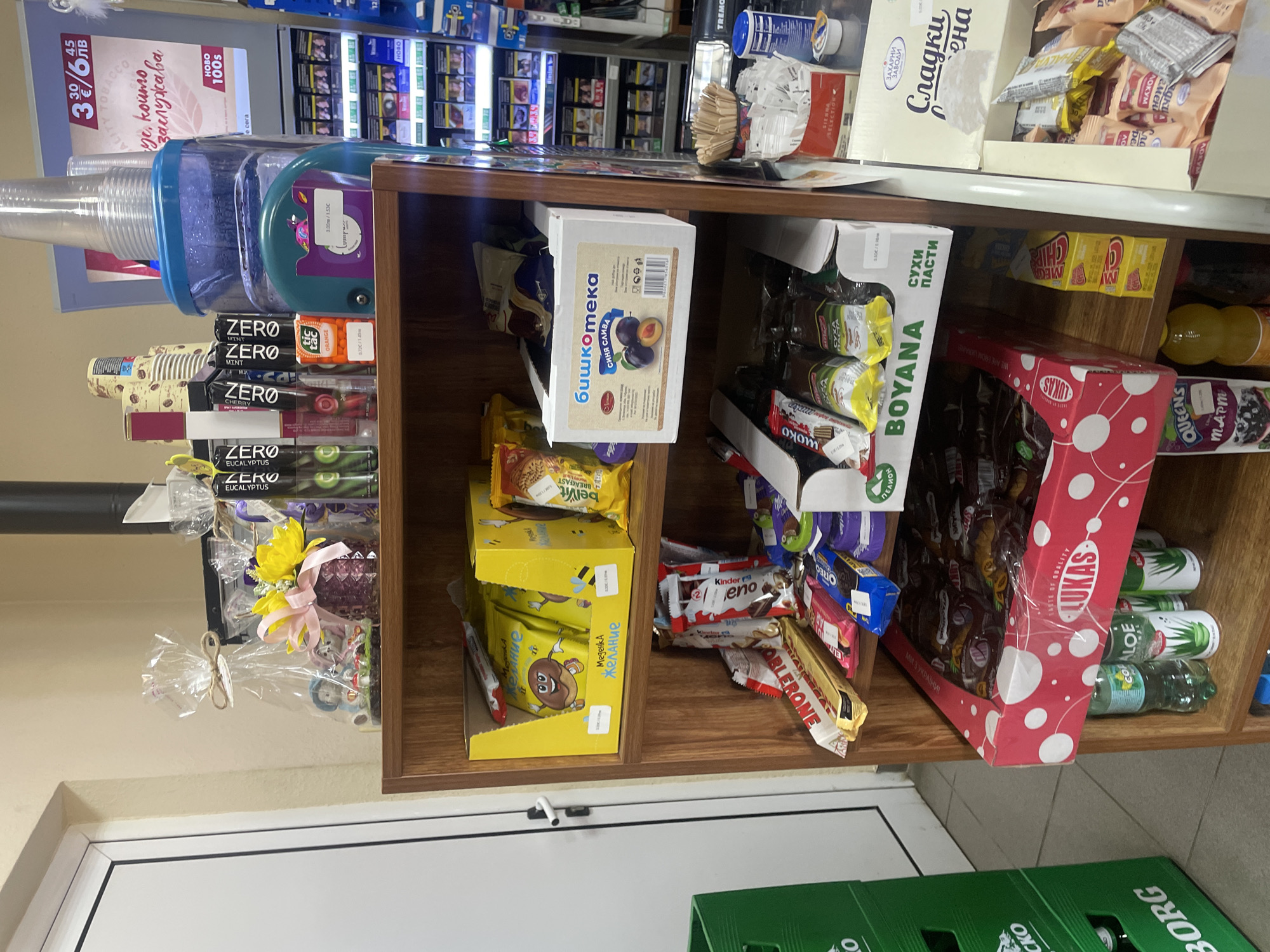 This image captures a section of an indoor retail setting, likely a convenience store or kiosk, located in Konstantinovo, Bulgaria. The scene is dominated by a three-tiered wooden display shelf unit in the foreground, packed with various consumer goods, primarily snacks, beverages, and confectionery items. No individuals are visible. On the top surface of the wooden shelf unit, from left to right, are: a clear cellophane-wrapped gift package tied with twine, containing small figurines; a dark purple patterned vase holding yellow flowers adorned with a pink ribbon; a stack of disposable paper coffee cups; several slim, black-packaged "ZERO" branded items in flavors like Eucalyptus and Mint; an orange Tic Tac box; a large clear plastic water pitcher with a blue lid, filled with water and topped with several inverted clear plastic disposable cups; a teal-colored plastic item with a sticker displaying "3.02лв / 5.92лв" (Bulgarian Leva, indicating a price); and a small, white decorative ornament resembling a feather or angel. Behind these items, a black vertical pipe is visible. The top shelf of the wooden unit contains two yellow cardboard display boxes on the left, each filled with individually wrapped "Меденка Желание" (Medenka Zhelanie - Honey Cake Desire) branded snacks featuring a cartoon donut. Visible prices are "0.50лв / 0.89лв". To the right, a white cardboard box labeled "Бишкотека синя слива" (Biskoteka Sini Sliva - Plum Biscuits), featuring a picture of plums, contains wrapped biscuits. A price of "3.00лв / 5.88лв" is partially visible. Other wrapped snacks are present behind these main displays. The middle shelf is filled with an assortment of individually wrapped chocolate bars and candies. Prominent brands include Toblerone (gold packaging), Kinder Bueno (red and white packaging), Oreo (blue packaging), and Milka (purple packaging). A white cardboard display box with green text "BOYANA СУХИ ПАСТИ" (BOYANA Sukhi Pasti - Dry Pastries) holds numerous individually wrapped pastries. The bottom shelf features a large red cardboard display box with white polka dots, branded "LUKAS," containing many individually wrapped items, likely chocolate bars or wafers. Beneath this box and to its right, several beverage bottles are visible, including a green cola bottle, two bottles of "ALOE" drink, a yellow bottle (possibly juice), and a dark-colored bottle. To the right of the "LUKAS" box, a stack of small yellow boxes labeled "MEGA CHIP" is present. Further to the right, a white cardboard box from "ЗАХАРНИ ЗАВОДИ" (Zaharni Zavodi - Sugar Factories)