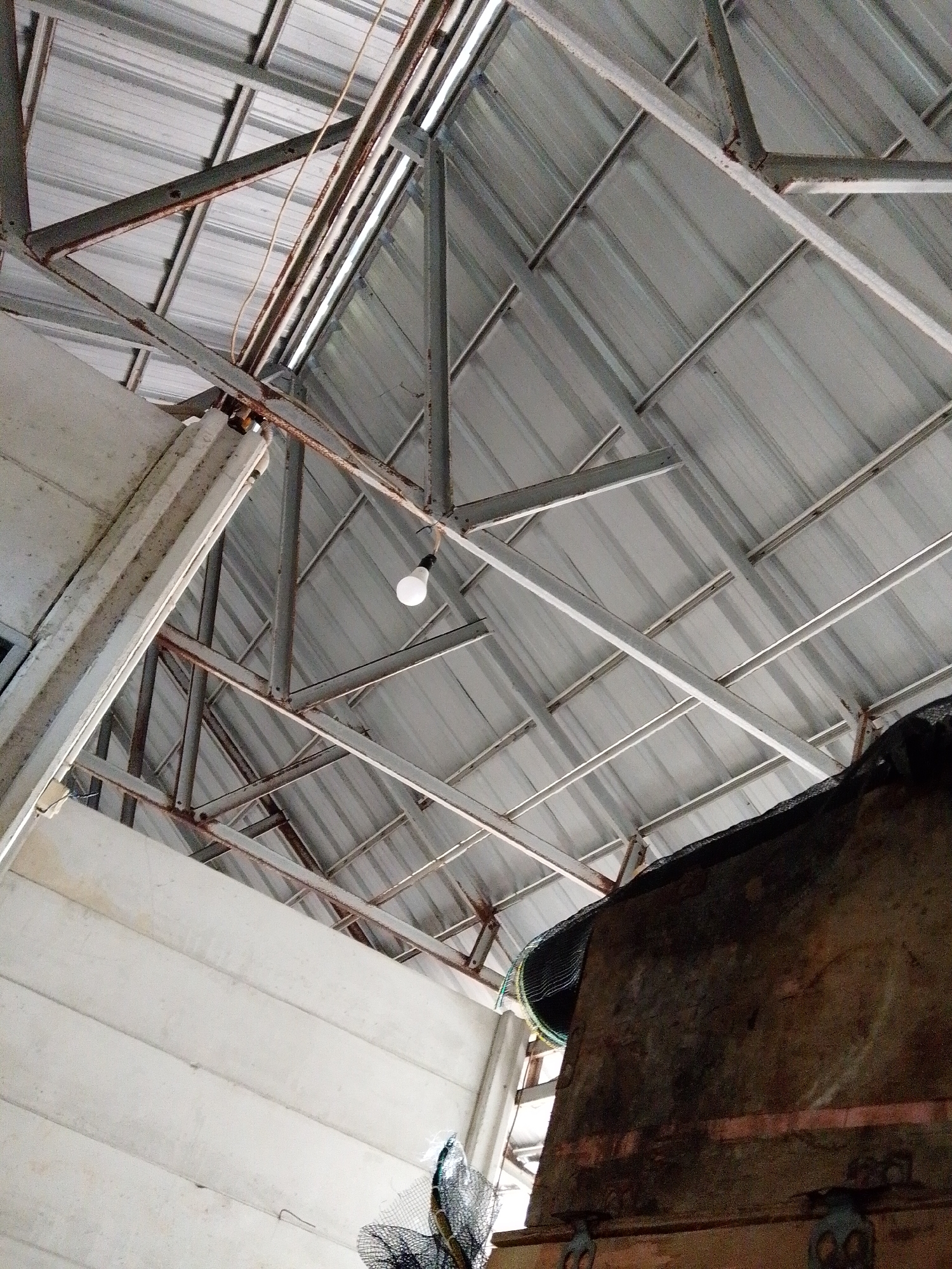 The image depicts the interior of a structure with a corrugated metal roof supported by a framework of metal beams. A single, bare lightbulb hangs from one of the beams. To the left, a section of white-painted wall is visible. A large, dark, wooden object occupies the lower right portion of the frame. The location is Bukit Ketok, Indonesia, but no additional details are available to support that assertion.