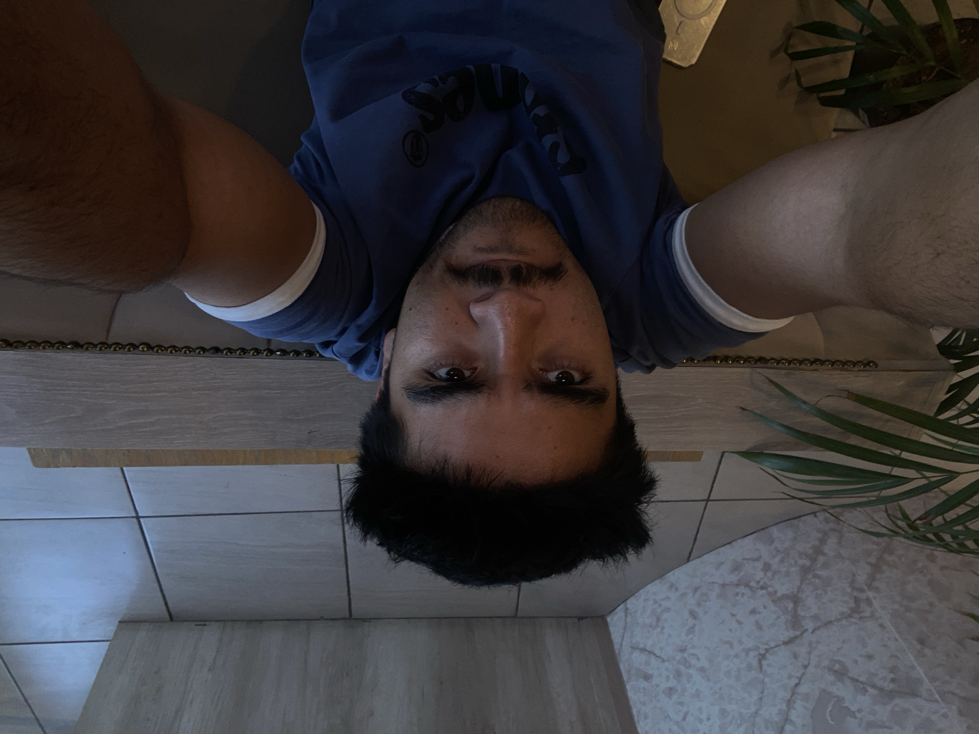 The image captures a close-up, low-angle self-portrait of a man, possibly taken while he is lying down and holding the camera overhead. The photo is rotated 90 degrees clockwise, so the man's head is positioned at the bottom of the frame and his body extends upwards.
The main subject is a man with short, dark hair, dark eyes, a mustache, and stubble. He looks directly into the camera with a neutral expression. He is wearing a dark blue t-shirt with white banding around the sleeves. Inverted text, possibly "JONES," is visible across the chest of his shirt. His forearms are visible on either side of his head, suggesting he is holding the camera.
The background reveals an indoor setting. Directly below his head (at the very bottom of the image) is a light-colored tiled floor, featuring a mix of plain and marble-patterned tiles with visible grout lines. Above his head and extending behind his shoulders is a wooden or laminate surface, possibly a table or bench, adorned with a decorative metallic strip composed of small, gold-colored spherical studs. To the far right of the frame, parts of a potted plant with slender green leaves are visible. The lighting is subdued, typical of an indoor environment, possibly in the evening. This scene is located in Puebla, Mexico.