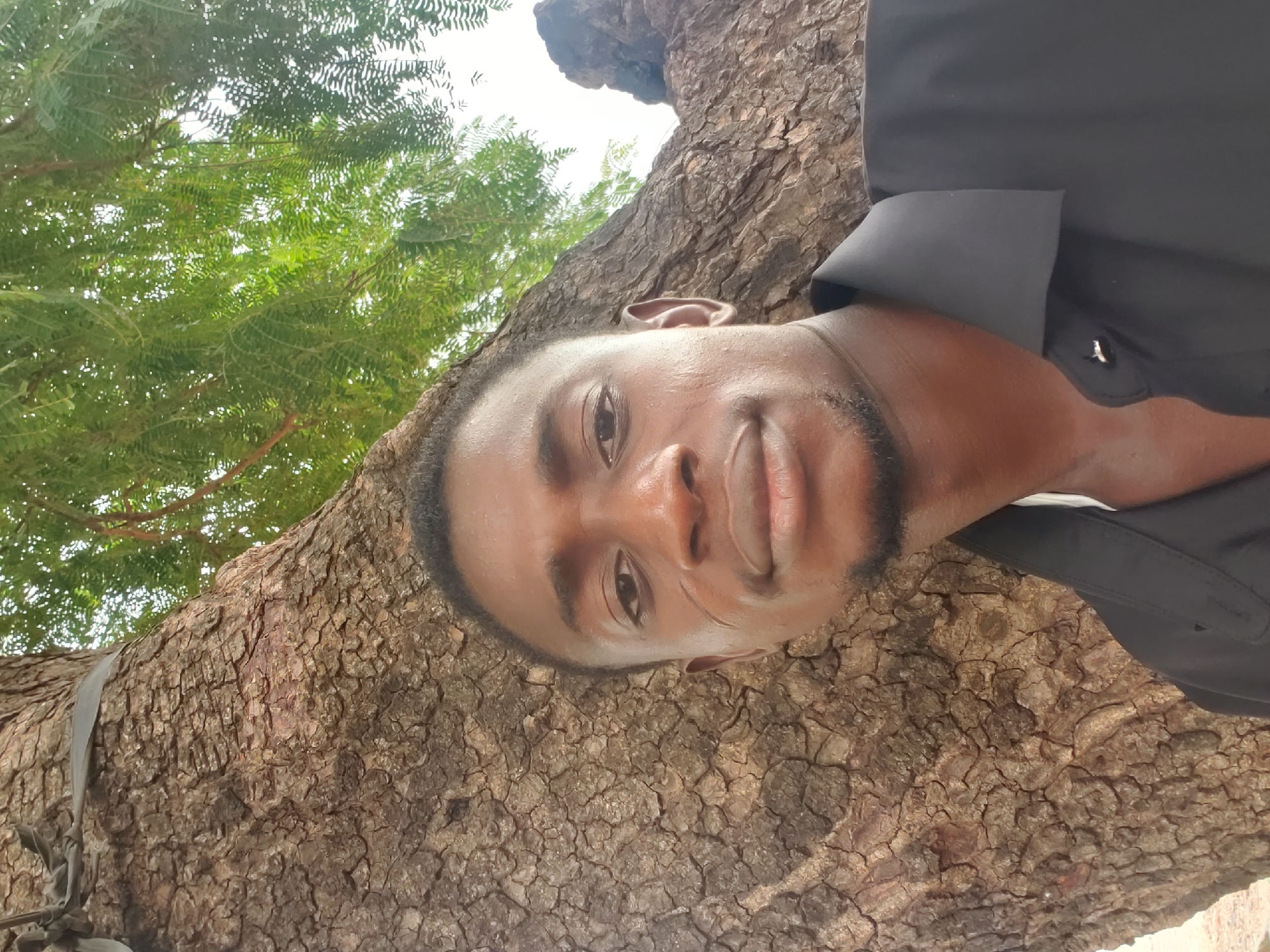A close-up, portrait-style photograph captures a young adult man positioned against the textured bark of a large tree. The image is taken at a slight upward angle, showing the man's face and upper torso. He has dark skin, short dark hair, and a short beard. He is looking directly at the camera with a calm, pleasant expression and a subtle smile. He is wearing a dark, collared shirt. A slight sheen on his face suggests warm or humid conditions.
The man is the central subject, with the rugged, deeply fissured bark of a large tree trunk forming the immediate background and extending across much of the lower and right portions of the frame. Above and to the left of the man, vibrant green foliage from the tree's canopy is visible against a bright sky, indicating an outdoor setting during the daytime. A dark strap or tie is visible wrapped around the tree trunk in the lower left corner. The setting is likely in Adankolo, Nigeria, given the context. No visible text is present in the image.