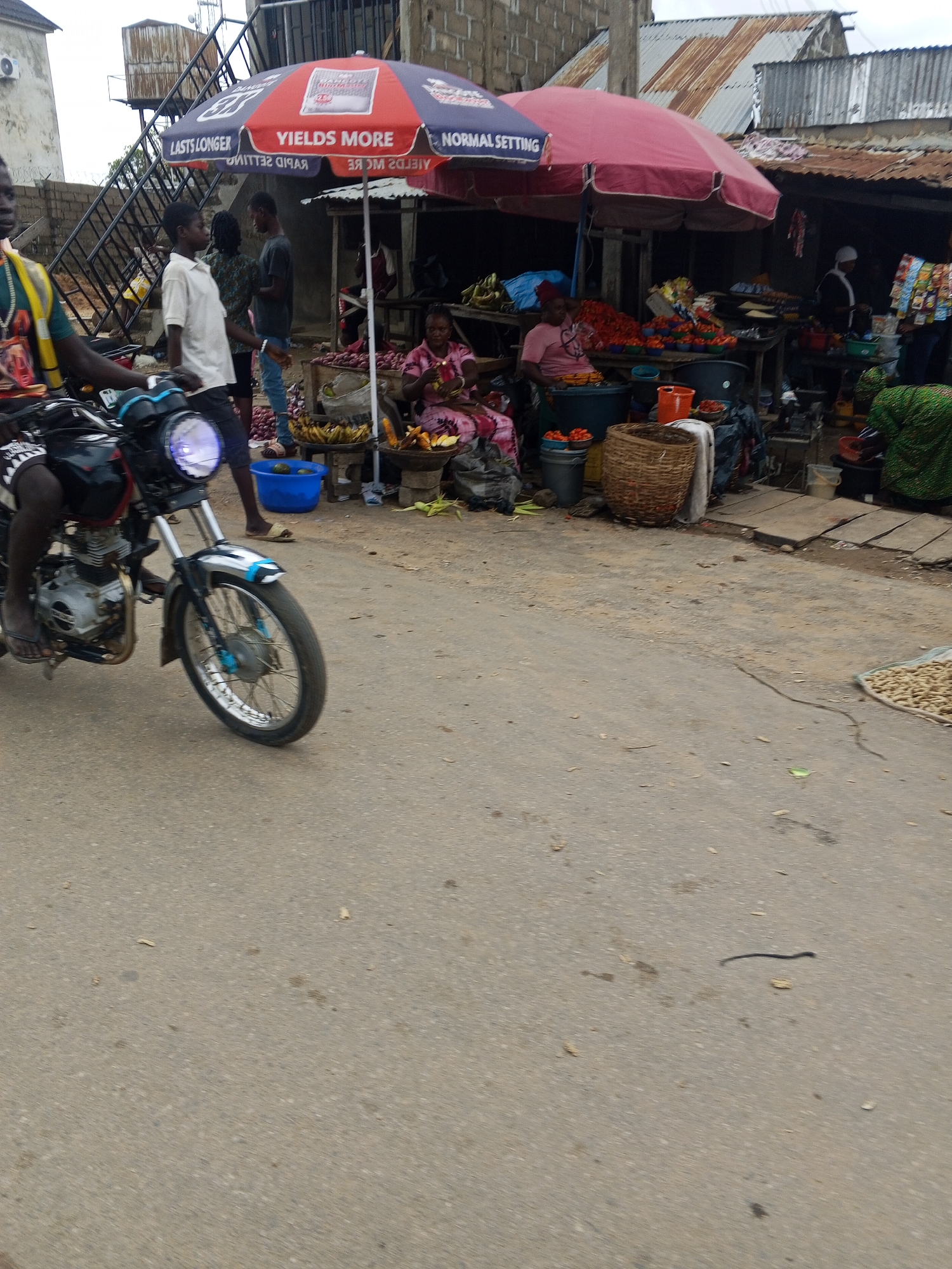 The image depicts a bustling outdoor market scene in Zango-ndaji, Nigeria. A person is riding a motorcycle in the foreground, its headlight illuminated. Behind the motorcycle, a market stall is set up under a large red and blue umbrella, displaying various produce including red peppers and bananas. Several people are visible at the market, some attending to stalls, and others browsing. A woman in a pink dress is seated near the produce. In the background, buildings with corrugated metal roofs are visible, along with a staircase structure. The ground is paved with asphalt, showing signs of wear and scattered debris. The overall atmosphere suggests a typical day in a Nigerian market, with activity and commerce underway.