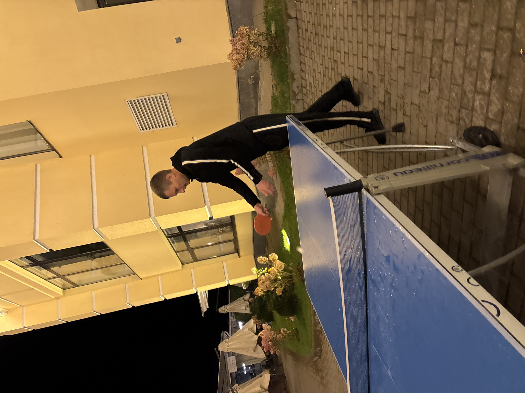 A man is playing table tennis outdoors at night. He is wearing a black tracksuit with white stripes and is holding an orange paddle. The table tennis table is blue and appears wet, suggesting it may have rained. The setting is a paved area next to a yellow building. In the background, there are several outdoor umbrellas and some flowering bushes. The scene suggests a recreational activity taking place in a public or residential area.
