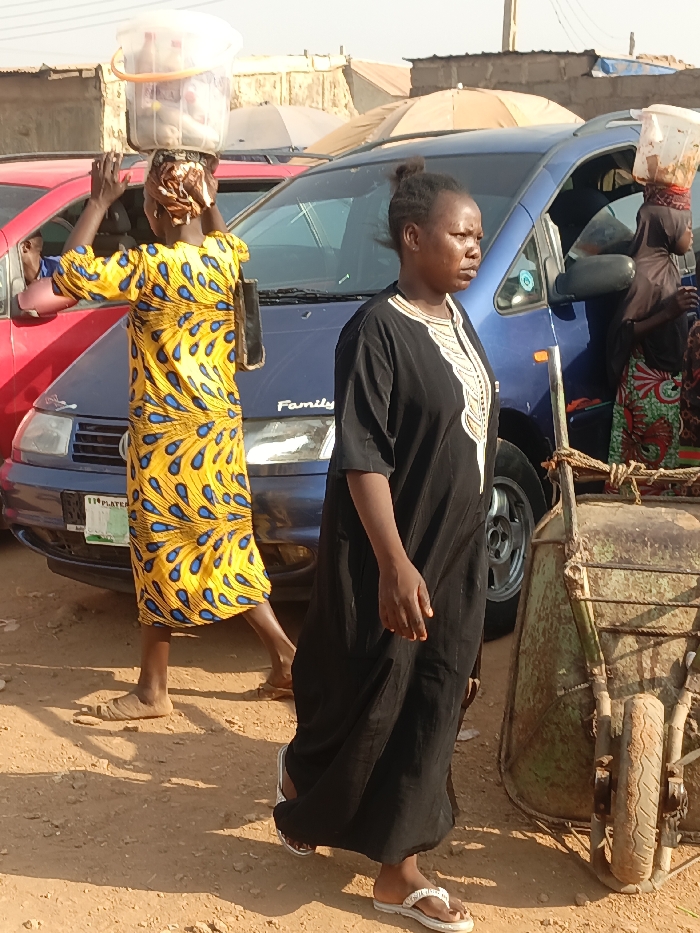 Two women are walking in an open dirt area. One is wearing a long black robe with intricate white embroidery around the neck and is facing forward. The other is wearing a long, bright yellow dress with a blue peacock feather pattern and is carrying a white bucket on her head. There are several cars parked in the background, including a red car on the left, and a blue van with the word "Family" on the back. Part of a green wheelbarrow is visible to the right.