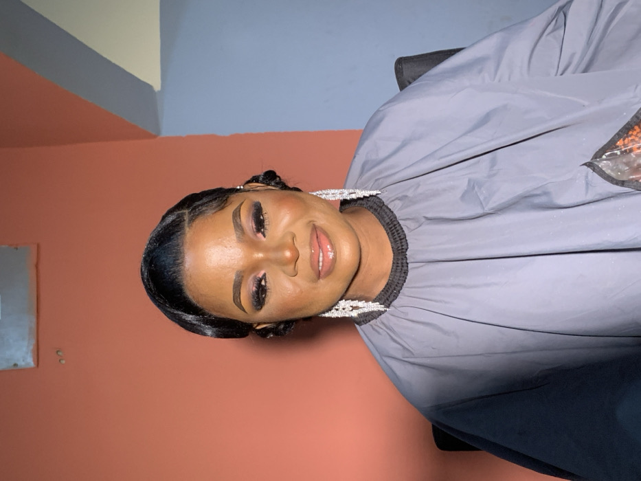 An adult female is centrally depicted in an indoor setting within the city of Garaku, Nigeria. She possesses dark skin, dark hair styled in sleek waves with a side bun detail, and expertly applied makeup including dark eyeshadow, eyeliner, and sculpted eyebrows, with glossy lips. She is wearing prominent, multi-tiered silver-toned chandelier earrings. Her torso is covered by a grey smock with a black elasticated collar, partially obscuring a dark chair. A translucent bag containing small orange items rests on the lower right section of the smock. The background consists of a wall painted reddish-orange on the left and light blue on the right, separated by an interior corner. An indistinct rectangular object and two small dark marks are present on the upper left wall. The subject maintains a subtle smile and faces directly forward.
