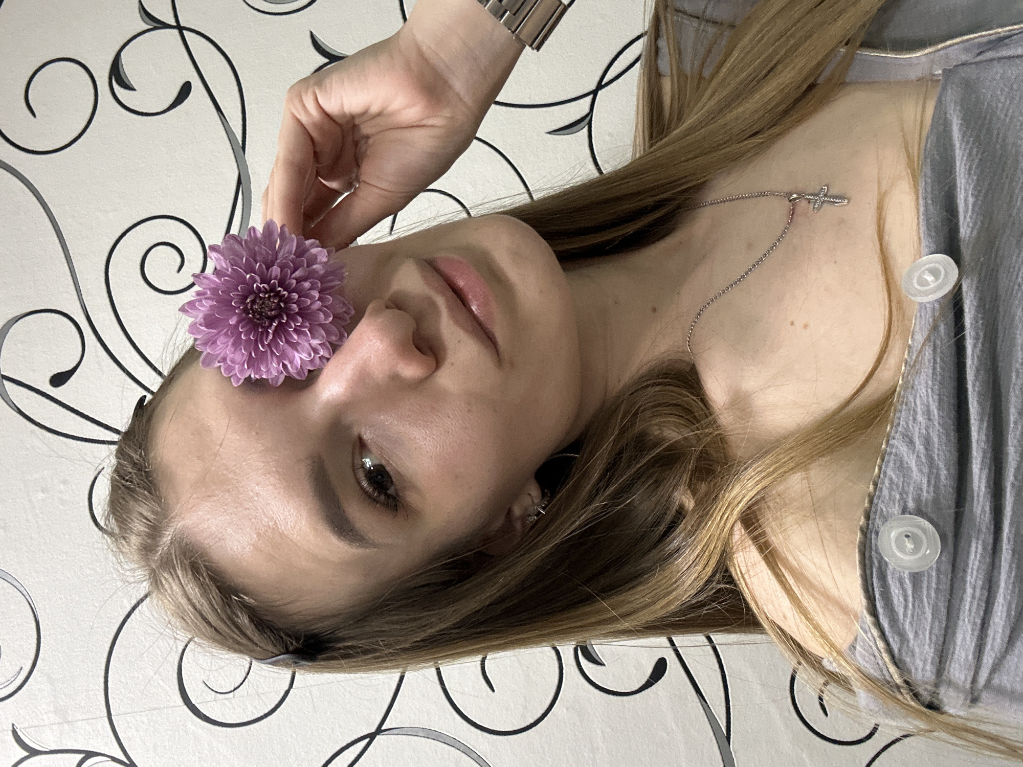 A woman with long, blonde hair lies against a white wall decorated with black and gray swirling patterns. She is wearing a gray shirt with white buttons and a silver necklace with a cross pendant. Her left hand is raised, holding a purple flower near her left eye. She appears to be looking towards the camera.