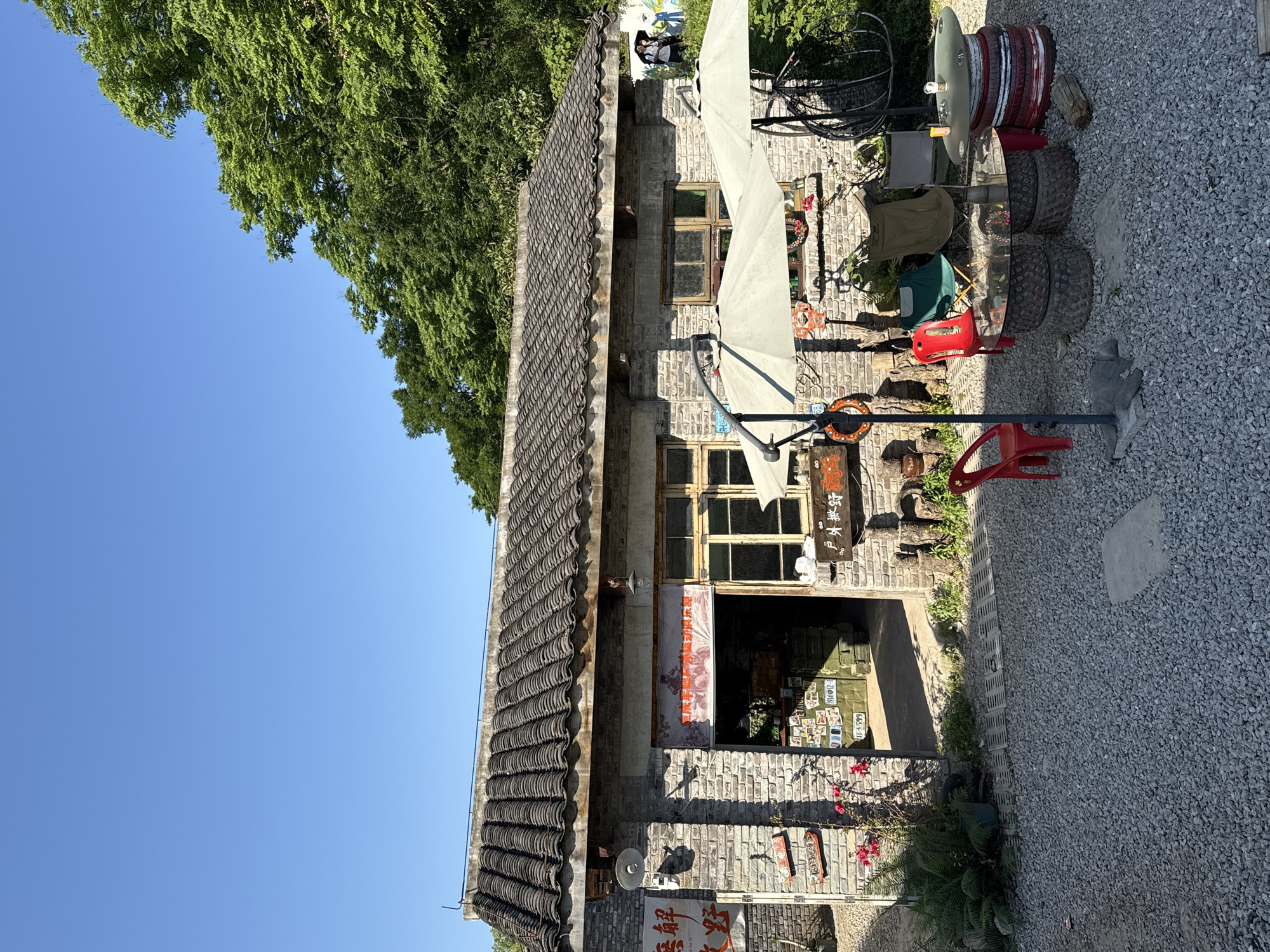 The image shows the exterior of a stone building with a dark tile roof. The sky is a clear blue, and green trees partially obscure the upper-left corner of the building. An umbrella is positioned in front of the building. Various objects, including tires and chairs, are scattered on the gravel ground next to the building. According to an online source, this may be the Old Factory Creative Park in Beibei, China.