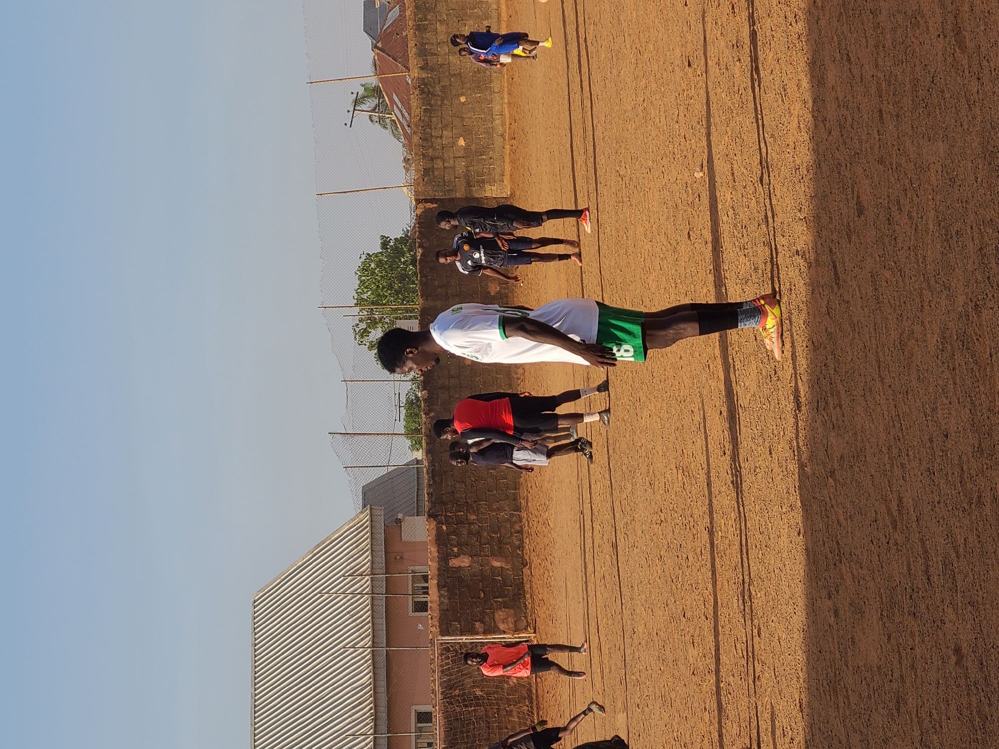 The image depicts an outdoor scene on a reddish-brown dirt field likely used for sports. Several people are present, possibly athletes engaging in warm-up exercises or drills. The person in the foreground is wearing a white jersey with green accents, black shorts, and colorful athletic shoes. Other individuals are wearing various athletic outfits, including red shirts and blue uniforms. A chain-link fence runs along the background, behind which are buildings with corrugated metal roofs and brick walls. The location is suggested to be in Jos, Nigeria, though no definitive landmarks are apparent. The sky is clear, indicating sunny weather.