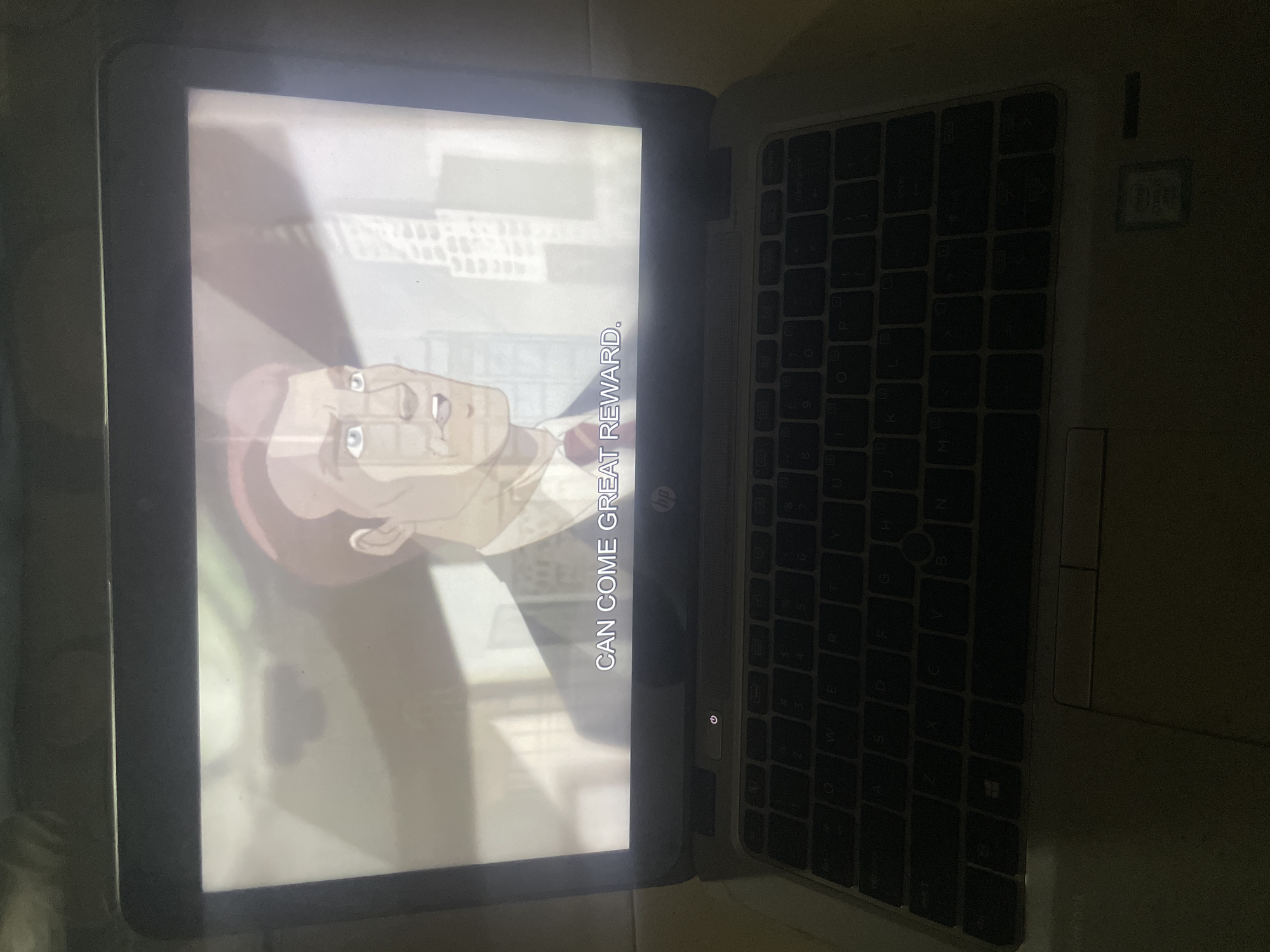 An animated character is displayed on the screen of a laptop computer. The character, a man with reddish-brown hair and light blue eyes, is wearing a suit and tie. Subtitles at the bottom of the screen read "CAN COME GREAT REWARD." The laptop is open, and the keyboard is visible. The screen exhibits glare and light reflections, suggesting the presence of ambient light in the surrounding environment. The laptop appears to be on a flat surface, though the surface is not clearly discernible. The image's composition and lighting suggest it was taken in a dimly lit indoor setting. No specific location in Ihiagwa, Nigeria, can be identified from the provided image.