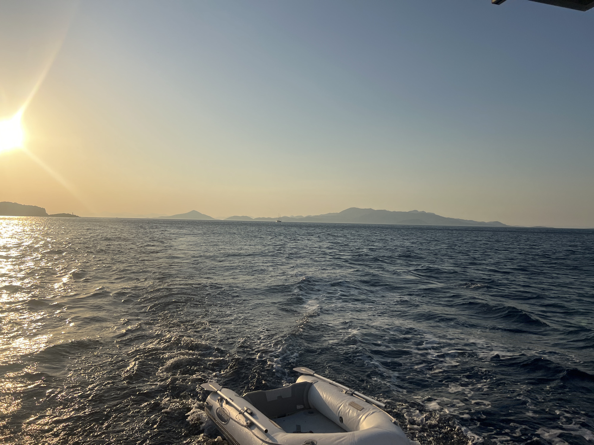 A wide shot shows an inflatable dinghy with oars being towed behind a larger vessel, creating a foamy wake in the dark blue sea. The sun, low on the left horizon, casts a golden sheen on the water and illuminates the sky with a soft yellow and blue gradient. In the distance, a hazy silhouette of mountains forms a landmass across the horizon. The image is taken from the perspective of someone on the towing vessel, looking out at the open water.