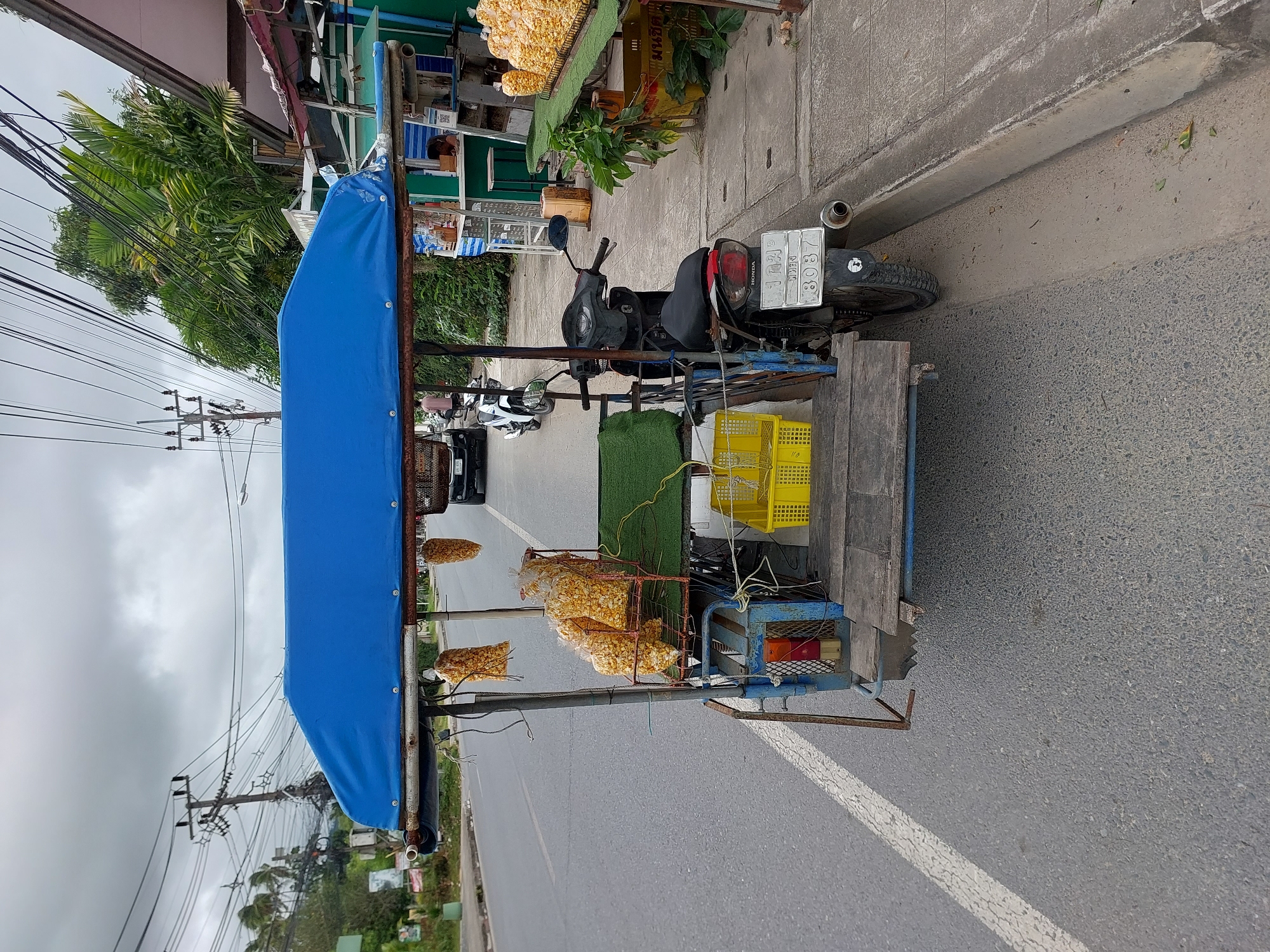 This image depicts a street scene in the Ko Samui District of Thailand. A blue-canopied cart, attached to a motorcycle, is parked on the side of a paved road. The cart is laden with bags of what appears to be popcorn or corn snacks, some of which are hanging from the canopy. There is a yellow plastic basket on the cart, and a green mat. The motorcycle is parked in front of the cart, and has a license plate visible with the number "8037". In the background, there are power lines, palm trees, and a building that appears to be a shop or stall, where a person can be seen. The sky is overcast. The overall scene suggests a roadside vendor setup.