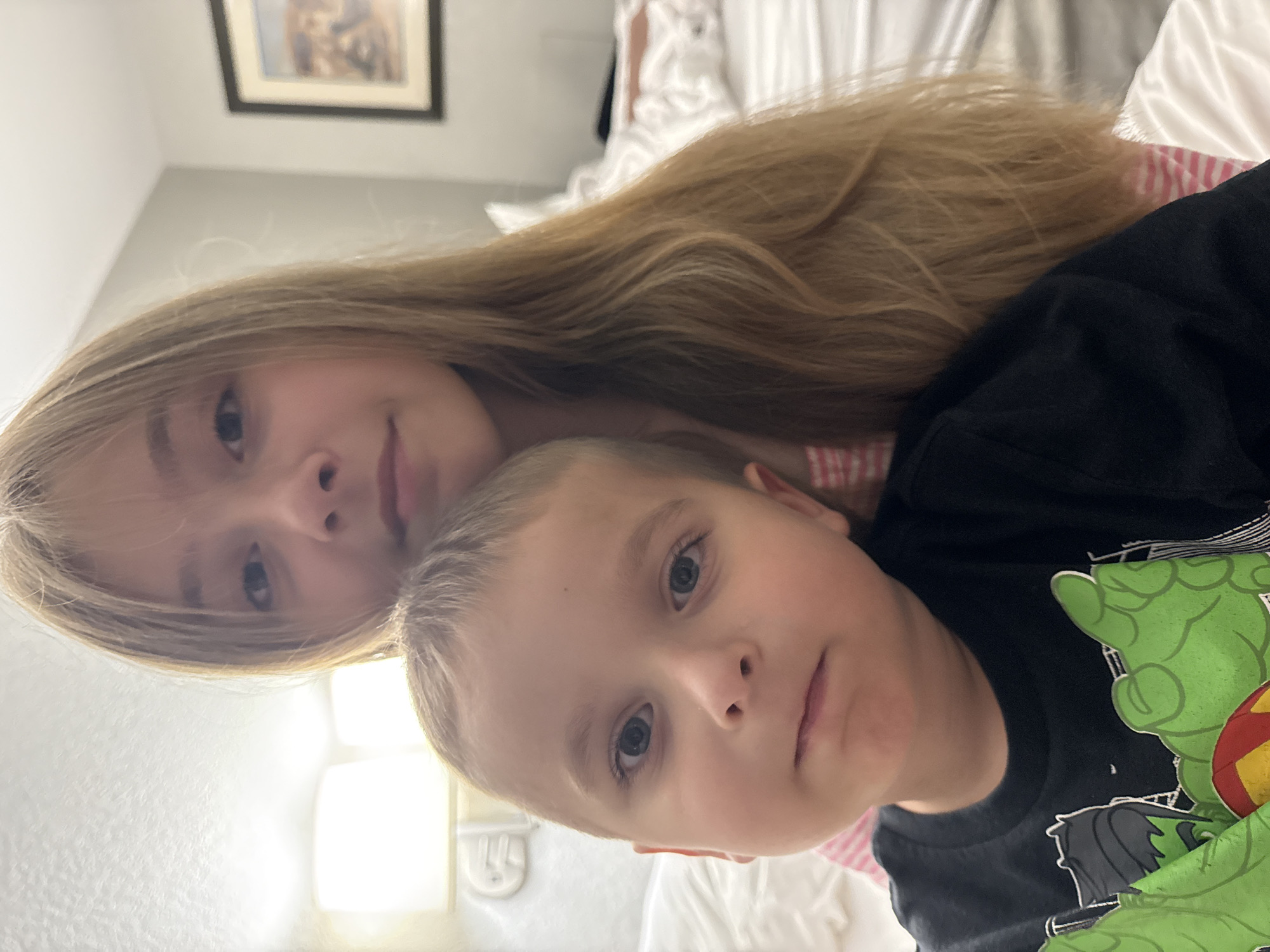 This is a close-up, indoor, eye-level, portrait-style photograph of two young children. A blonde girl with long, wavy hair is behind a younger blonde boy. The boy, in the foreground, is wearing a black t-shirt with a green Hulk graphic. He is looking at the camera with a neutral expression. The girl, in the background, is wearing a pink and white striped shirt and is also looking at the camera. Her hair falls over her shoulders. The background appears to be a hotel room, with a bed and a framed picture on the wall. The photograph was taken in Myrtle Beach, United States of America.