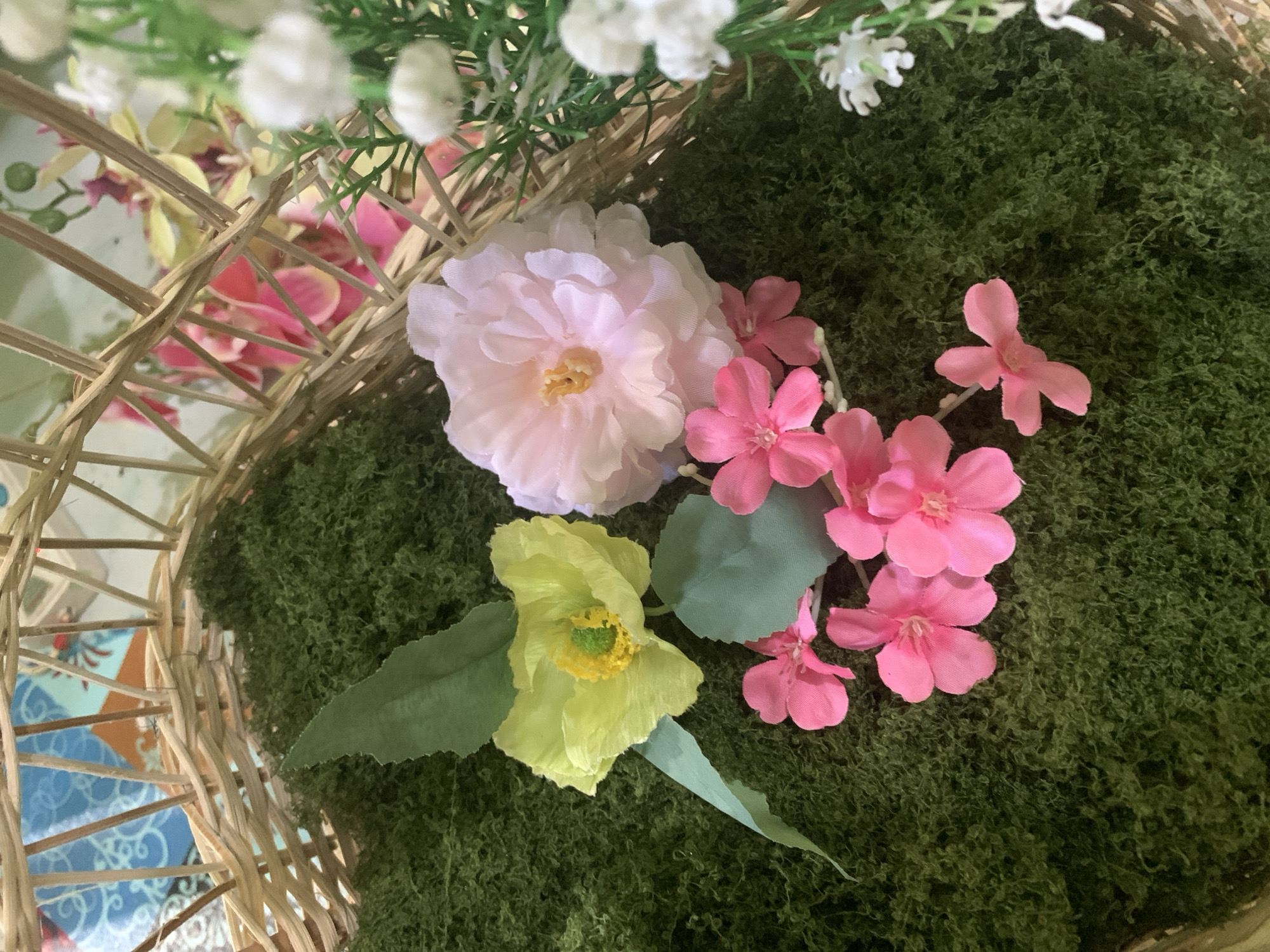 The image shows a close-up view of a wicker basket filled with artificial moss and flowers. A pale yellow poppy-like flower with a yellow and green center sits on the left, accompanied by two light green leaves. To its right is a larger, pale pink flower with many delicate petals. A cluster of smaller, bright pink flowers with five petals each is arranged below these larger blooms. In the upper right portion of the image, out of focus, are sprigs of small white flowers and green foliage. The background behind the basket reveals a pattern of blue and orange fabric, and glimpses of more artificial flowers in pink and yellow hues. The setting does not appear to be in Kuala Pembuang, Indonesia, nor does it depict any recognizable urban landscape or specific geographic features. The scene is a still life arrangement of decorative floral elements.