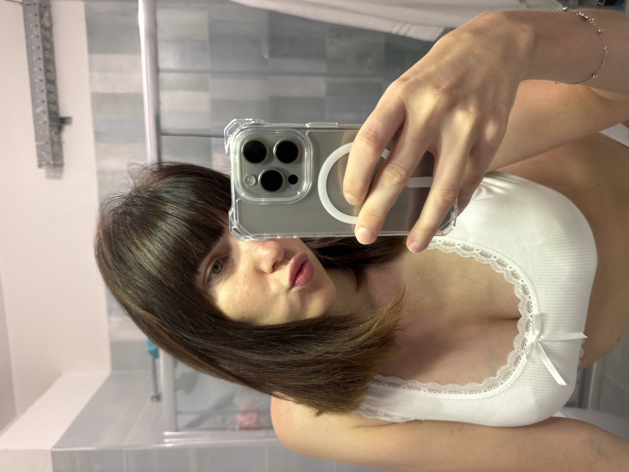 A young woman is taking a selfie in a bathroom mirror, likely in her home. She is wearing a white tank top with lace trim and a small bow at the center. Her hair is brown and shoulder-length, with bangs. She is puckering her lips towards the camera. She is holding a clear phone case with a camera array. The bathroom appears to be tiled with pale blue and white tiles, and there is a shower stall and a towel rack visible in the background. The woman's left arm is visible, wearing a delicate silver bracelet with small beads.