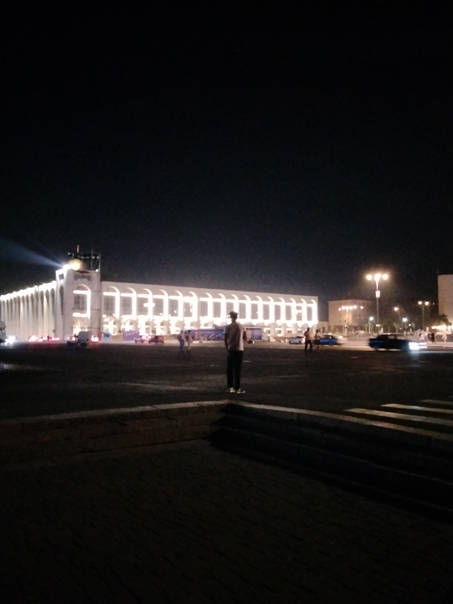 The image depicts an exterior night scene, possibly a public square or plaza, in Bishkek City, Kyrgyzstan. In the foreground, a lone individual stands with their back to the camera, facing a large, illuminated building. The building, which appears to be a long, multi-arched structure, is brightly lit with white lights along its facade and features a prominent tower with a searchlight or spotlight projecting a beam into the night sky. Several other people and vehicles are visible in the mid-ground, near the illuminated building and on a road in front of it. Streetlights are also visible in the distance, contributing to the overall illumination of the scene. The ground in the foreground is paved with dark material and shows raised, tiered elements. The overall impression is one of a well-lit, modern urban environment at night.