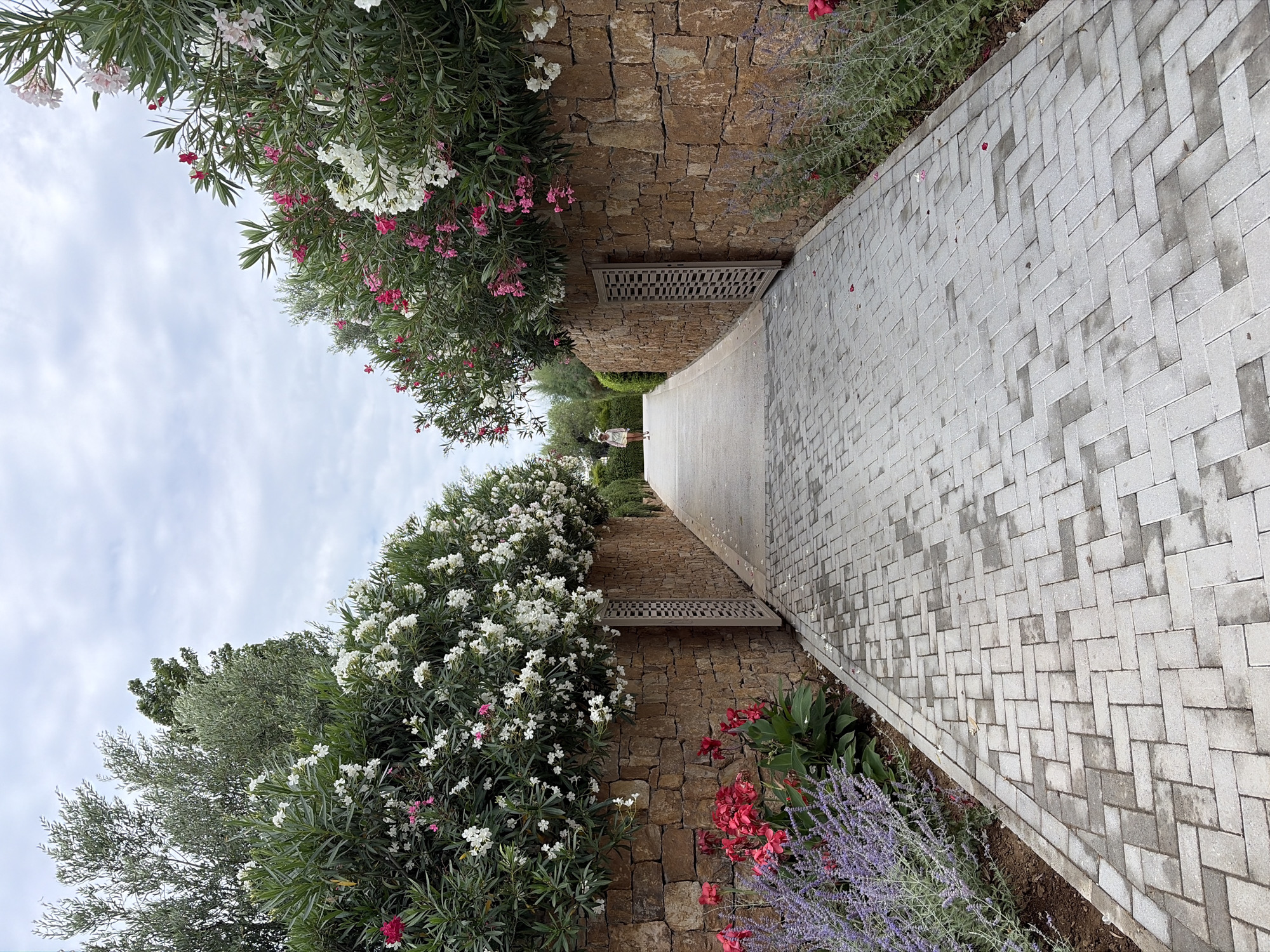 A paved pathway with light gray interlocking bricks leads into the distance. The pathway is flanked by rough-hewn stone walls. On the left, a person is walking away from the camera towards the end of the pathway. The walls are topped with lush greenery and flowering bushes, featuring white and pink blooms. Red and purple flowers are also visible along the left edge of the pathway, closer to the camera. On the right, the stone wall continues, with similar greenery and flowers above. The sky is overcast and cloudy. The scene is an outdoor setting, likely a garden or a walkway in a resort area within the Kassandra Municipal Unit, Greece, characterized by its Mediterranean flora and stonework.