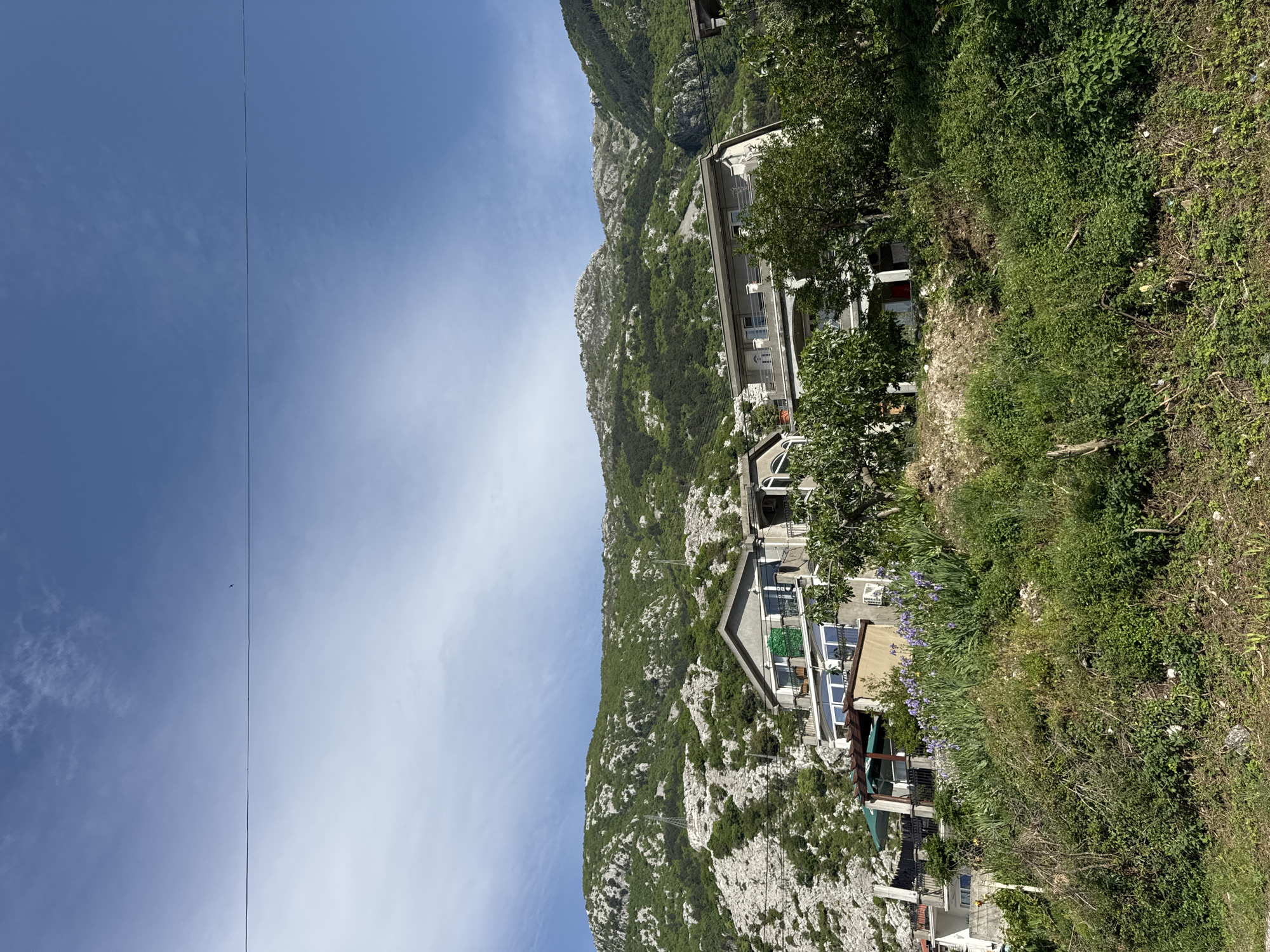 The image captures a view of Škaljari, Montenegro, with a steep, rocky mountain partially covered in greenery in the background. The foreground features a few multi-story buildings built into the mountainside, interspersed with lush vegetation. An electrical cable extends across the sky. The sky is blue with scattered clouds.