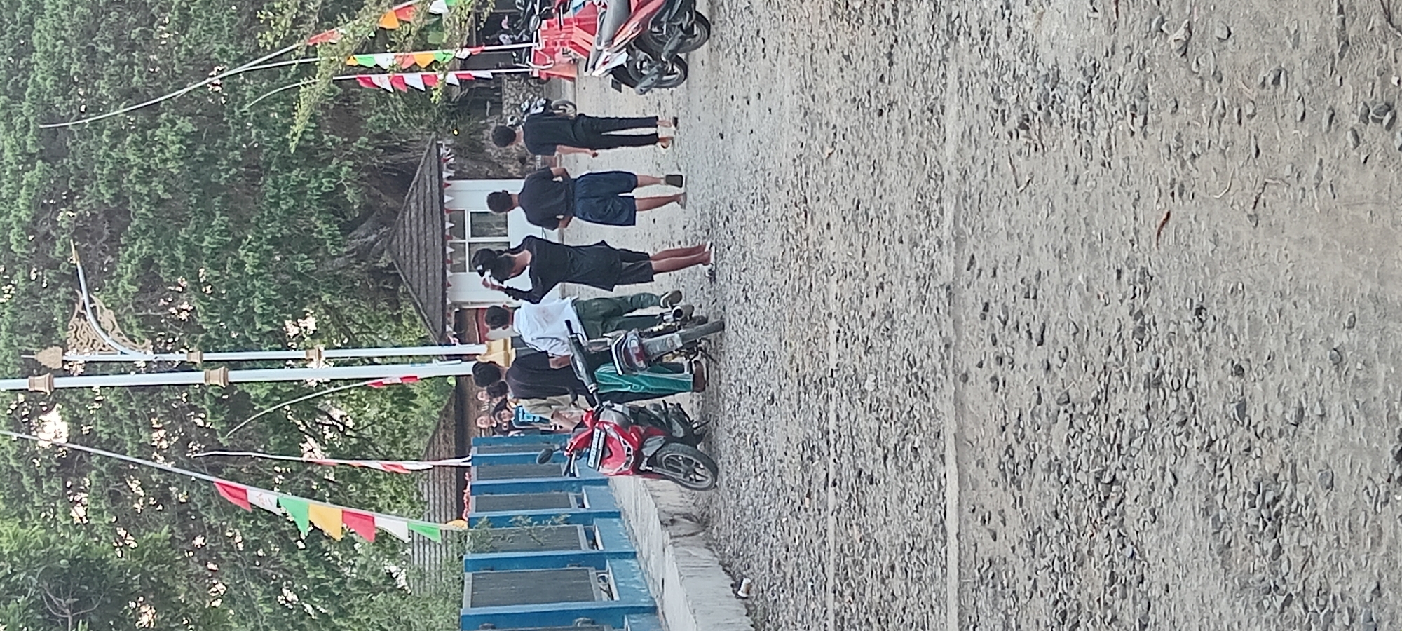 In the city of Kalipucang, Indonesia, on a gravel road with a blue-fenced structure to the left, a group of young men stand near two motorcycles. One motorcycle, a red scooter, is parked on the left. Another motorcycle, a darker scooter, is parked to its right. The men are dressed in casual clothing, with some wearing t-shirts and shorts, and one in a white long-sleeved shirt. One of the men appears to be adjusting his hat. In the background, there is a large tree, a small building with a dark roof, and several flag pennants strung between poles. The scene suggests a casual gathering or stop on the side of the road.
