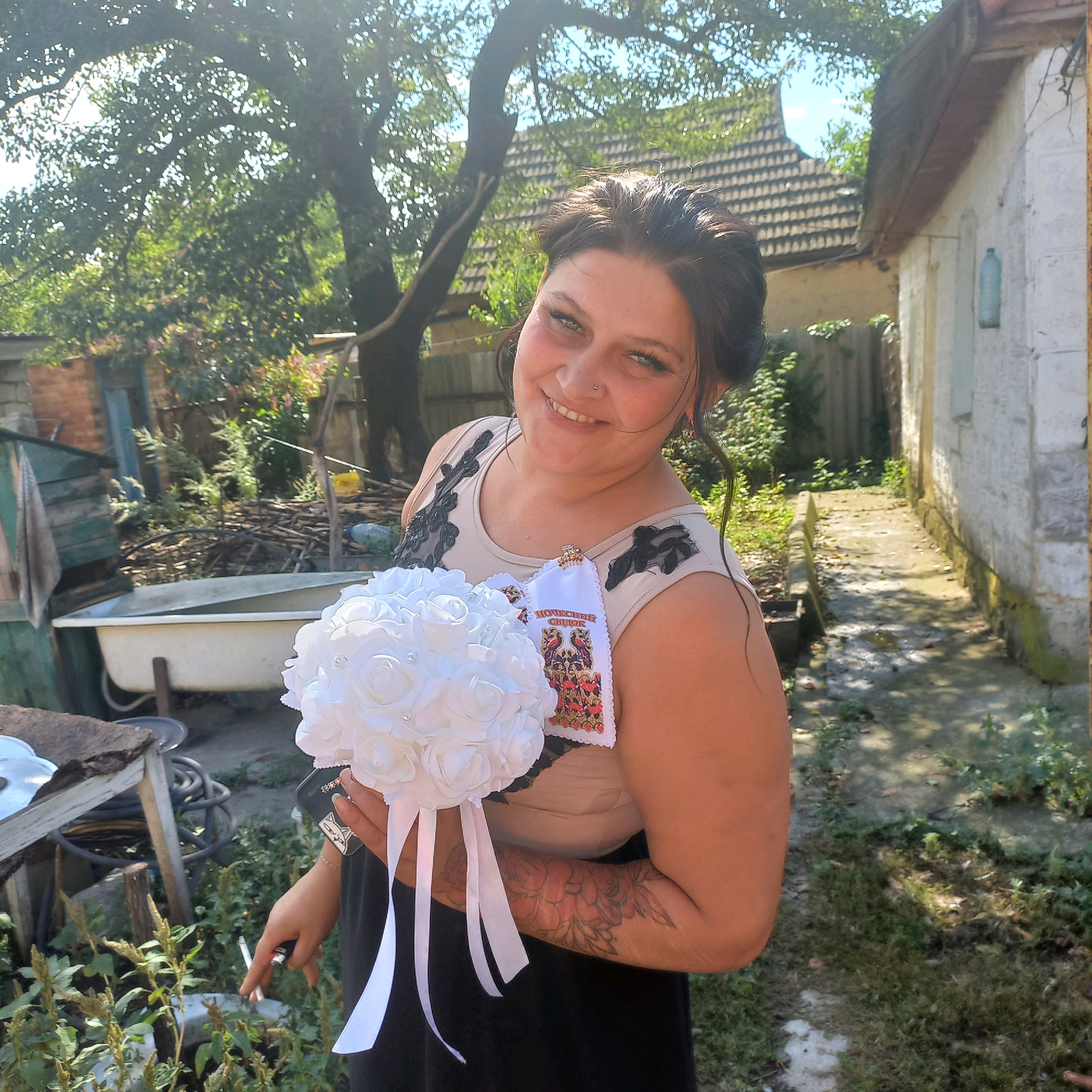 In Pokrovske, Ukraine, a woman stands outdoors in a sunlit yard, holding a bouquet of white roses. She is wearing a neutral-colored sleeveless top with black lace detailing and a dark skirt. A white embroidered cloth is pinned to her top, featuring a red and black design with birds. The background shows a residential area with houses, trees, and a bathtub in the yard. The woman is smiling at the camera and appears to be holding a lit cigarette. Her left arm is adorned with a floral tattoo. The overall impression is a candid moment captured in a rustic, outdoor setting.