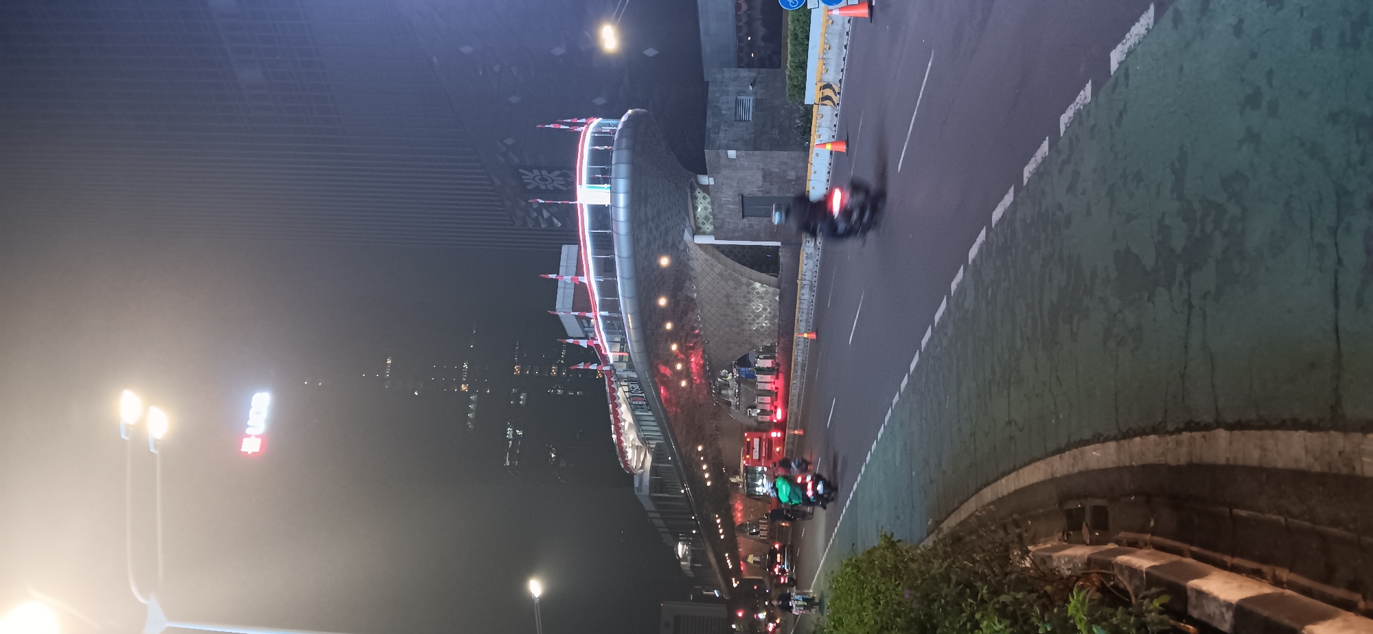 This image depicts a street scene at night in Central Jakarta, Indonesia, specifically near Bundaran HI. A modern building with a facade resembling a ship's bow is illuminated by red and white lights, with flags adorning its upper edge. The building appears to be an elevated pedestrian walkway or bridge. The street in the foreground is wet, suggesting recent rain or a humid night, and features traffic, including several motorcycles and a bus. The overall atmosphere is dimly lit, with streetlights casting a hazy glow and the background dominated by tall, modern buildings shrouded in mist or fog. The scene captures the dynamic urban environment of Jakarta after dark.