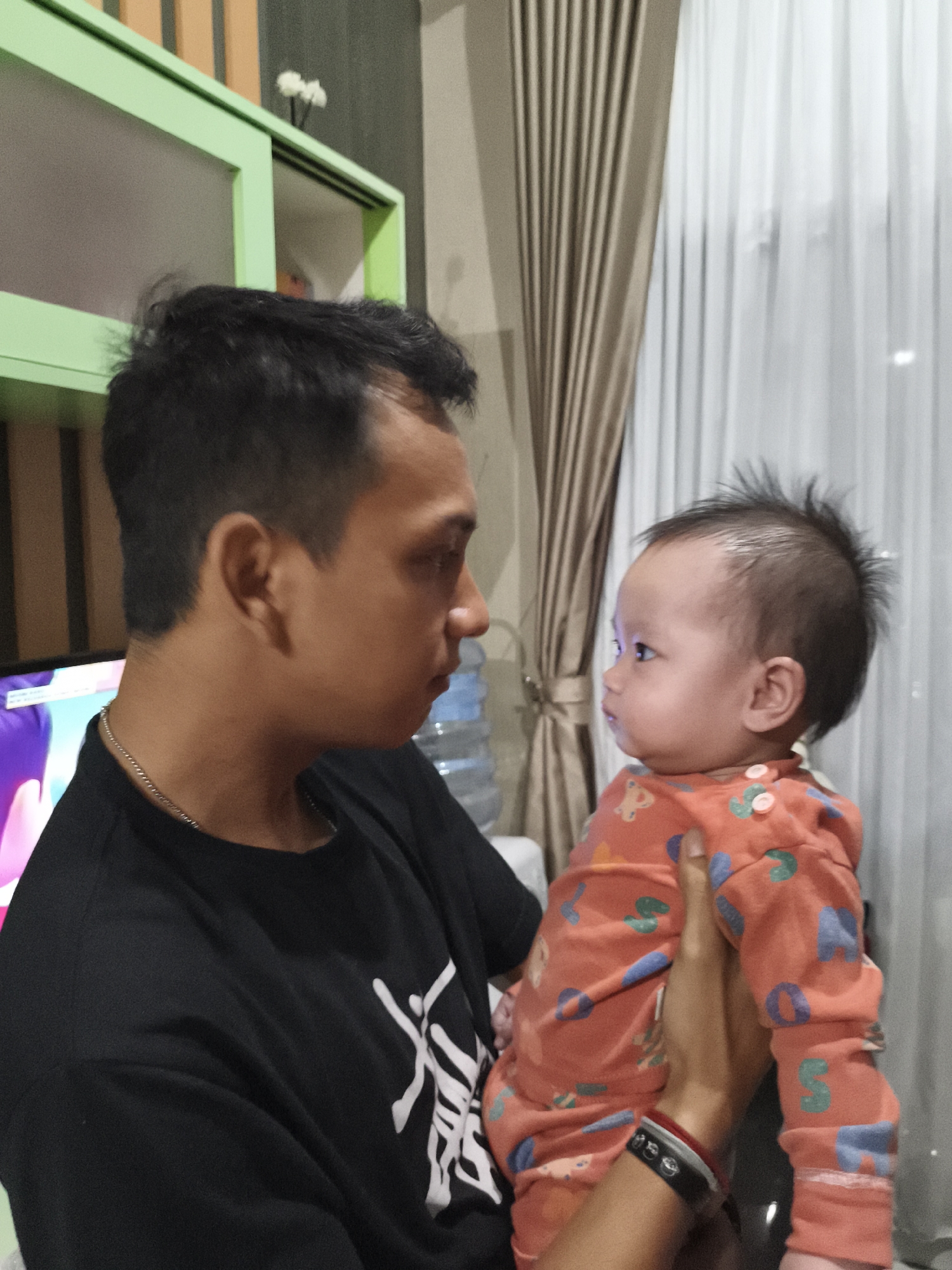 The image shows a man and a baby indoors, possibly in a home in Depok, Indonesia. The man, wearing a black t-shirt and a silver chain, is holding a baby. The baby, dressed in an orange sleepsuit with colorful patterns, is facing the man and looking towards him. Behind them, a light-colored curtain is visible, and to the left, a green cabinet and a computer monitor can be seen. The overall impression is a candid moment between a caregiver and a child in a domestic setting.