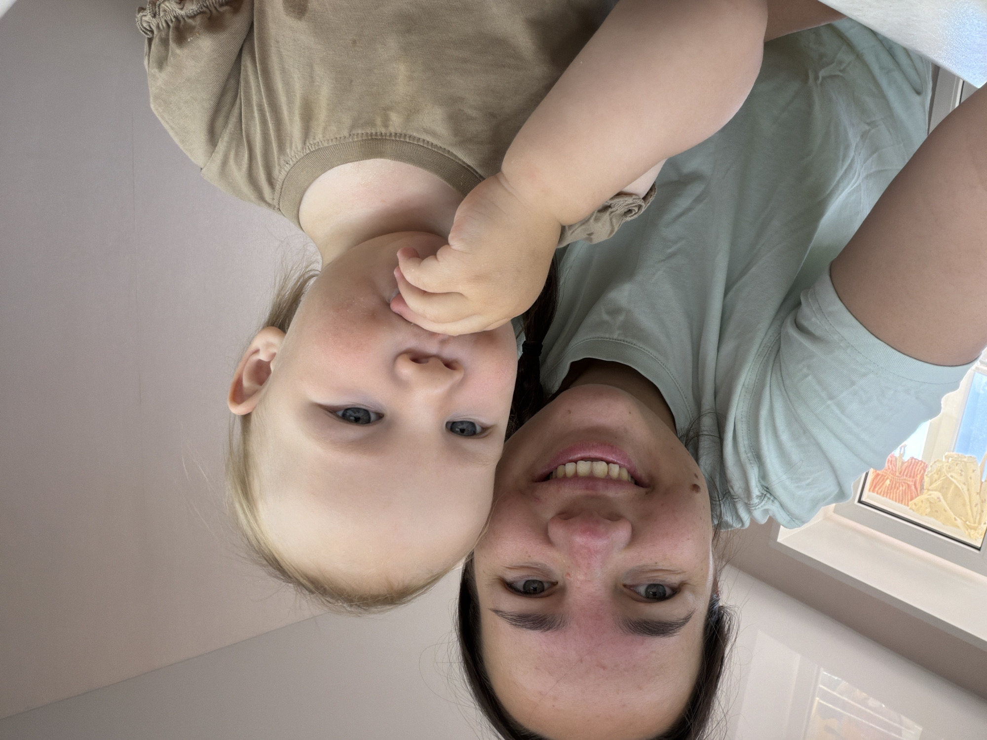 A woman and a toddler are taking a selfie in a room. The woman has dark hair and is wearing a light green t-shirt. The toddler has blonde hair and is wearing a tan t-shirt. The toddler is putting their hand in their mouth. The background appears to be a domestic interior, possibly a home, with a window visible in the left side of the frame showing laundry hanging outside. The overall lighting suggests daytime. The location is identified as Chaikovsky, Russia.