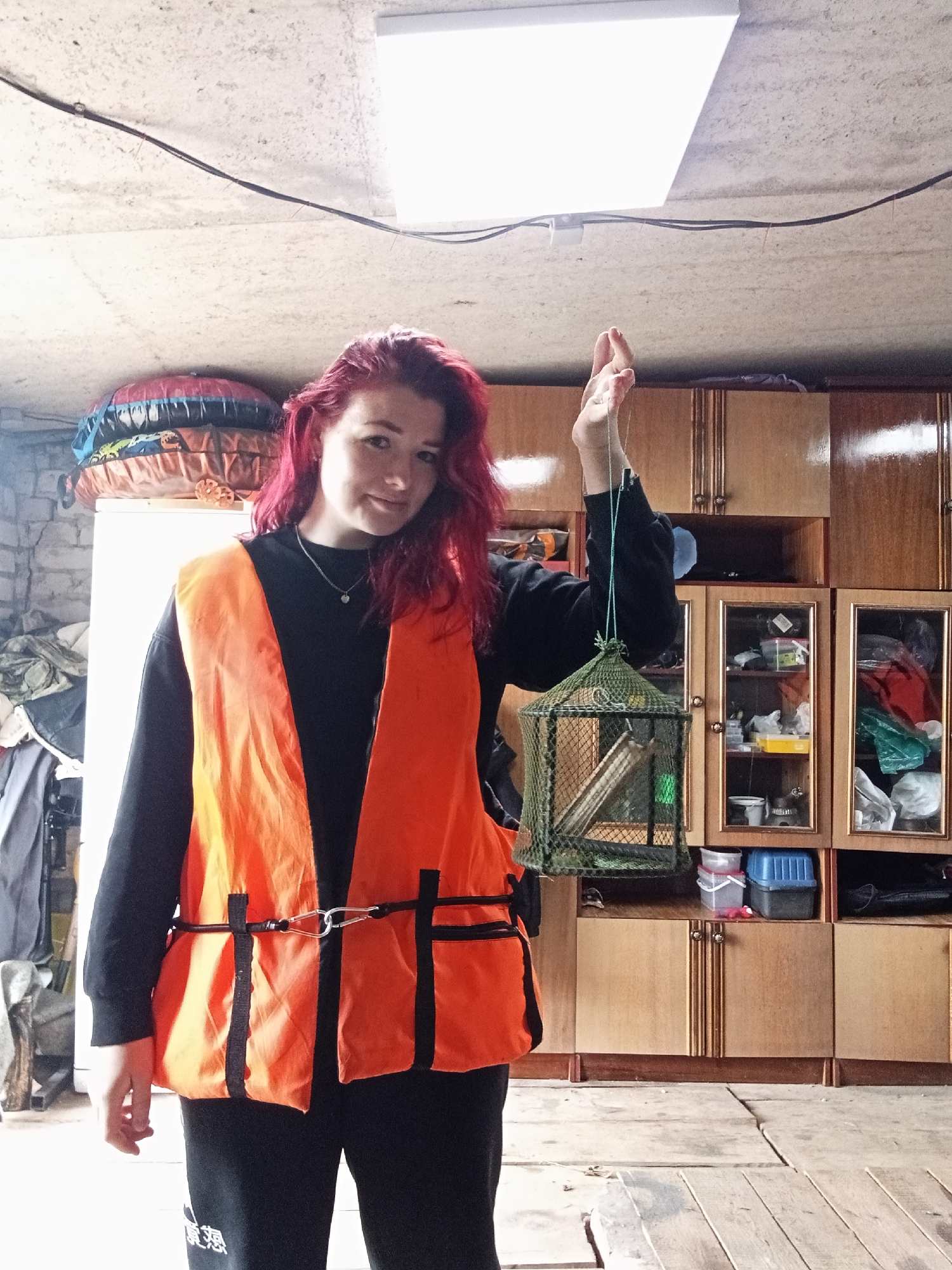 A young woman with bright pink hair stands in what appears to be a garage or storage area. She is wearing an orange safety vest over a black sweatshirt and black pants. In her right hand, she holds a small, green mesh cage suspended by a string. The cage contains what looks like some sort of bait or lure. Behind her, there are wooden cabinets filled with various items, including containers and what appear to be fishing supplies. The floor is made of rough wooden planks. The ceiling is concrete with exposed wires and a bright fluorescent light fixture. To the left of the woman, there are stacks of what seem to be inflatables or sleeping bags. The lighting suggests it is daytime, but the overall environment is dimly lit.