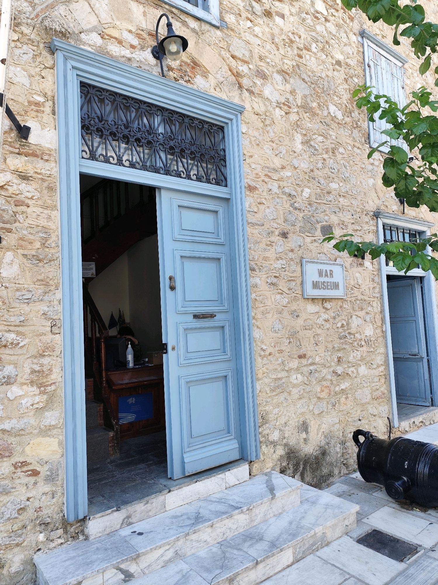 This is a full exterior shot of the War Museum in Nafplio, Greece. The museum is housed in a traditional stone building with a light blue door and window shutters. The building has a rough stone facade. The main entrance is an open, light blue door revealing a dark interior with a staircase. Above the door, there is an ornate wrought-iron grille. To the right of the entrance, a sign on the wall reads "WAR MUSEUM". A large, black cannon is positioned on the ground to the right of the entrance steps. The steps leading up to the entrance are made of marble. A tree with green leaves is visible on the right side of the image, partially obscuring a window.