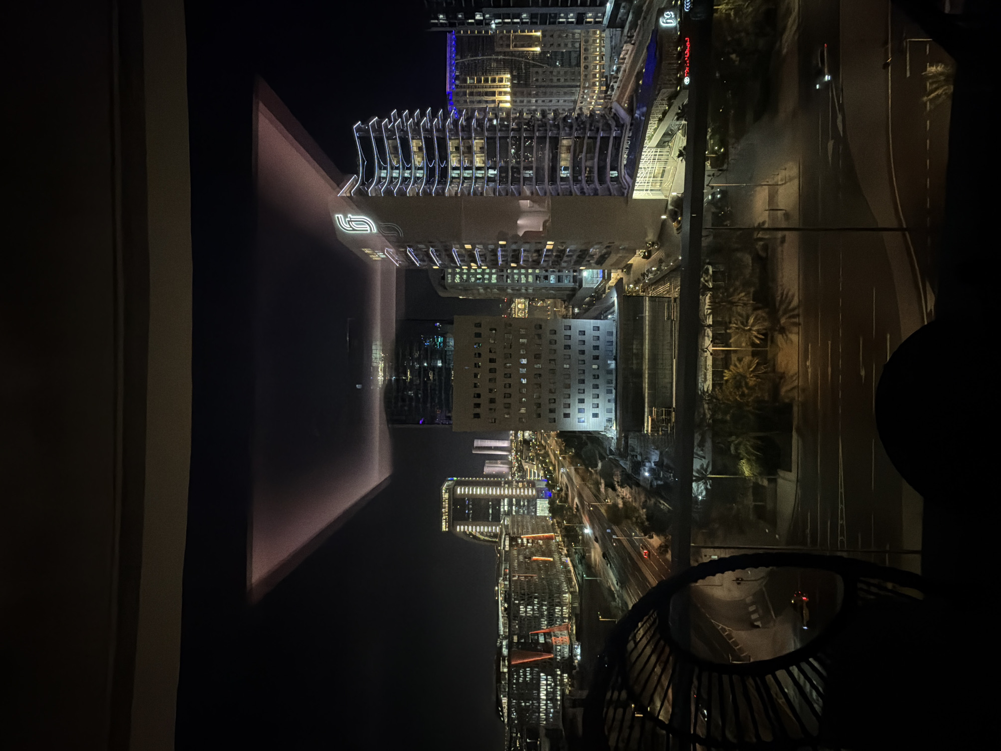 A night-time, elevated, medium shot captures a cityscape dominated by modern architecture and illuminated buildings. The foreground features a dark, curved structure with a circular opening, obscuring the bottom left of the frame. Beyond this, a wide road stretches into the distance, with visible streetlights and several cars, some with headlights on. The road is bordered by green spaces and palm trees, casting shadows.
The midground showcases a cluster of contemporary buildings of varying heights and designs. One prominent building on the right is tall and slender, adorned with horizontal white LED lighting along its edges and a stylized logo at its peak. Adjacent to it are other structures with multi-story windows and intricate architectural details. Further into the distance, a broad expanse of city lights stretches across the horizon, indicating a densely populated urban area.
Above the cityscape, a large, dark, angular structure with subtle pinkish-purple lighting is visible, its reflective surface adding a surreal quality to the scene. The sky is completely dark, suggesting nighttime. The overall impression is one of a technologically advanced and vibrant urban environment at night.