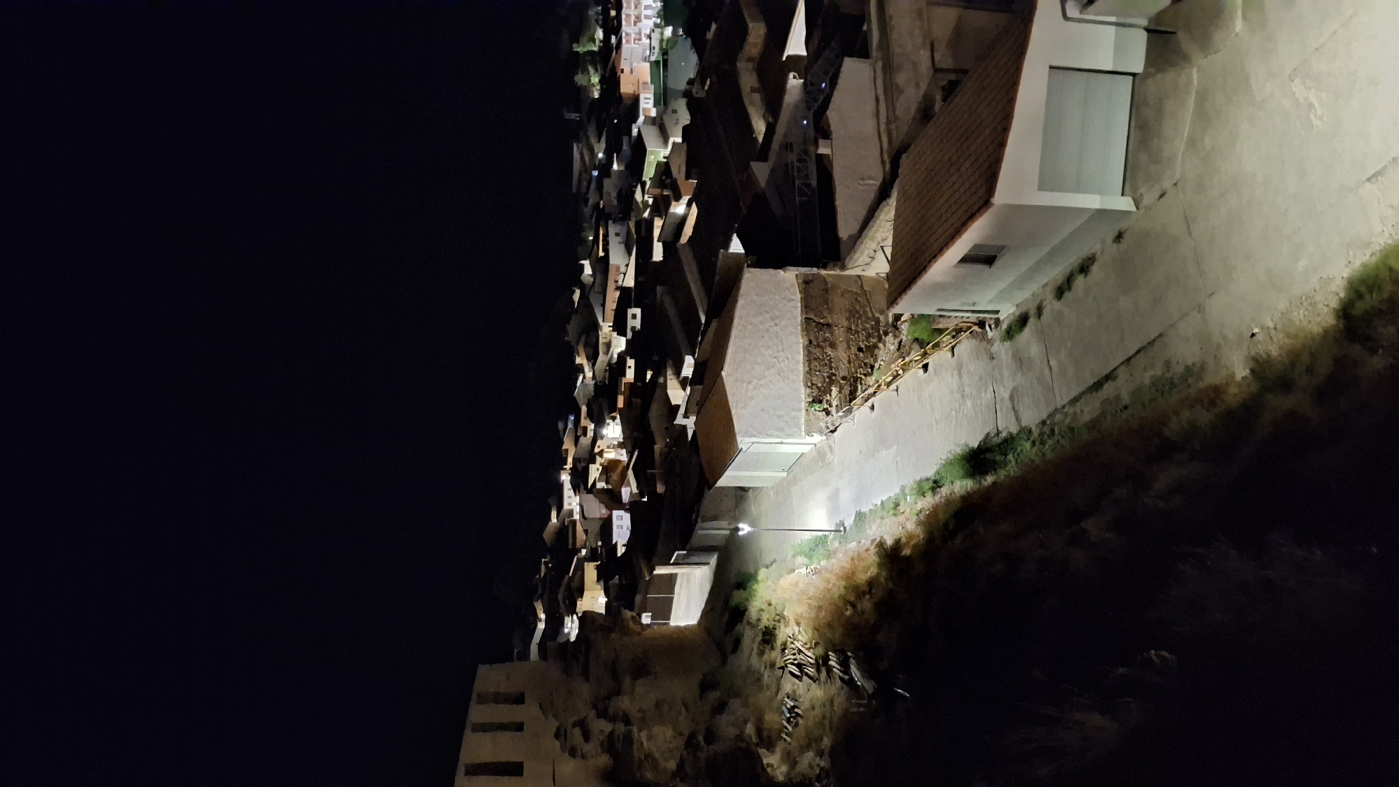 The image is a nighttime photograph overlooking a village in Híjar, Spain. A street lamp illuminates a path leading into the village, with houses and buildings constructed of stone and tile roofs stretching into the distance. On the left side of the frame, a rocky embankment with sparse vegetation rises, topped by the weathered facade of an old stone structure. The village is lit by a few scattered lights, suggesting it is either late at night or early in the morning. The overall impression is one of a quiet, traditional Spanish village under the cover of darkness.