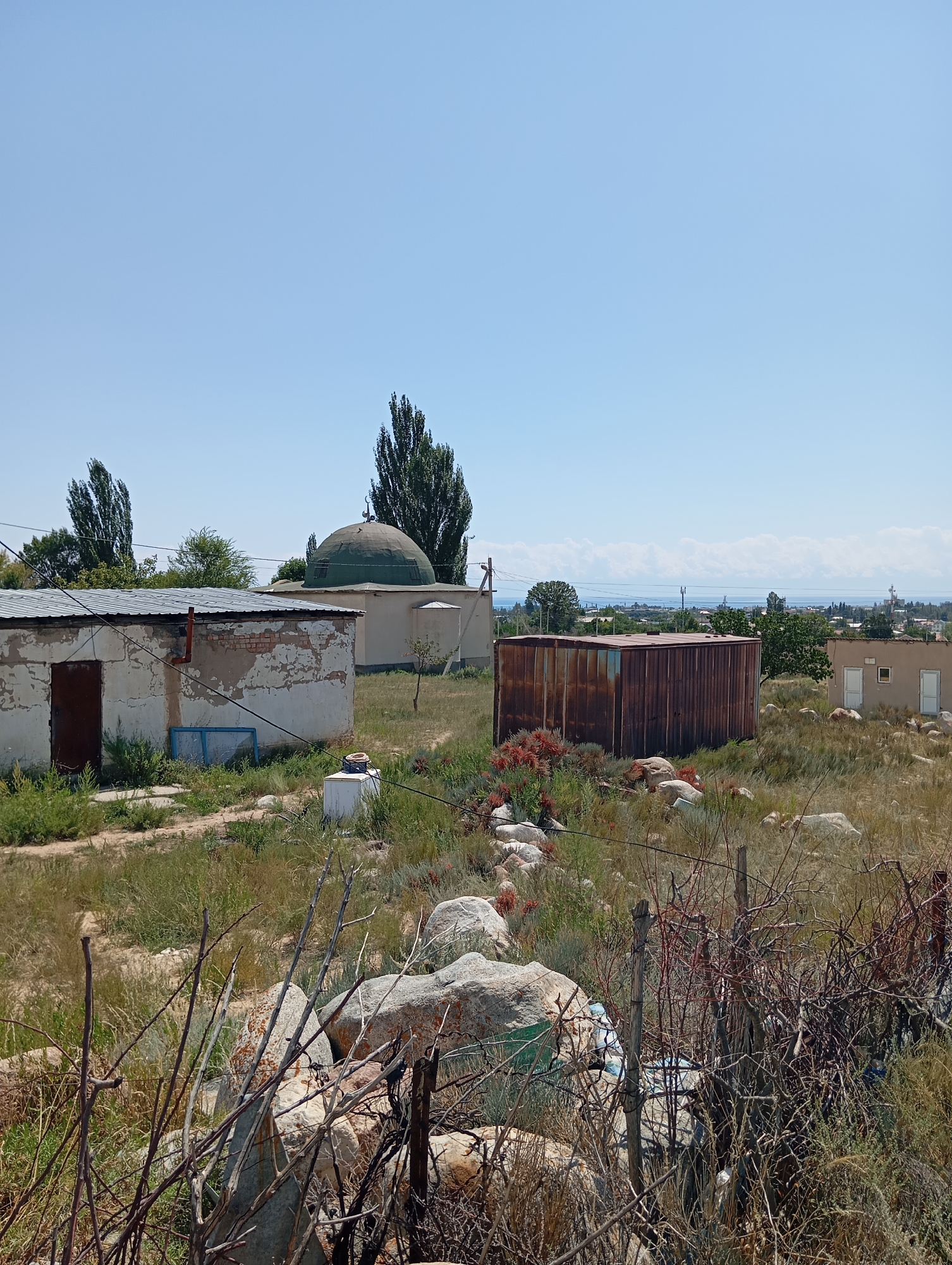 The image depicts a rural scene in Cholpon-Ata, Kyrgyzstan, under a clear blue sky. In the foreground, dry brush and large rocks are scattered across overgrown grass. A rusty, rectangular metal container, likely a storage unit, sits prominently in the middle ground. To its left, a dilapidated building with a corrugated metal roof and weathered, peeling walls exhibits a dark metal door. Further to the left, a green-domed structure, possibly a mosque or a mausoleum, is visible, partially obscured by trees. In the background, a distant body of water, likely Lake Issyk-Kul, is visible, with a hazy mountainous landscape beyond it. Several other smaller buildings and sparse vegetation are scattered throughout the landscape. The overall impression is one of a somewhat neglected, rural settlement.