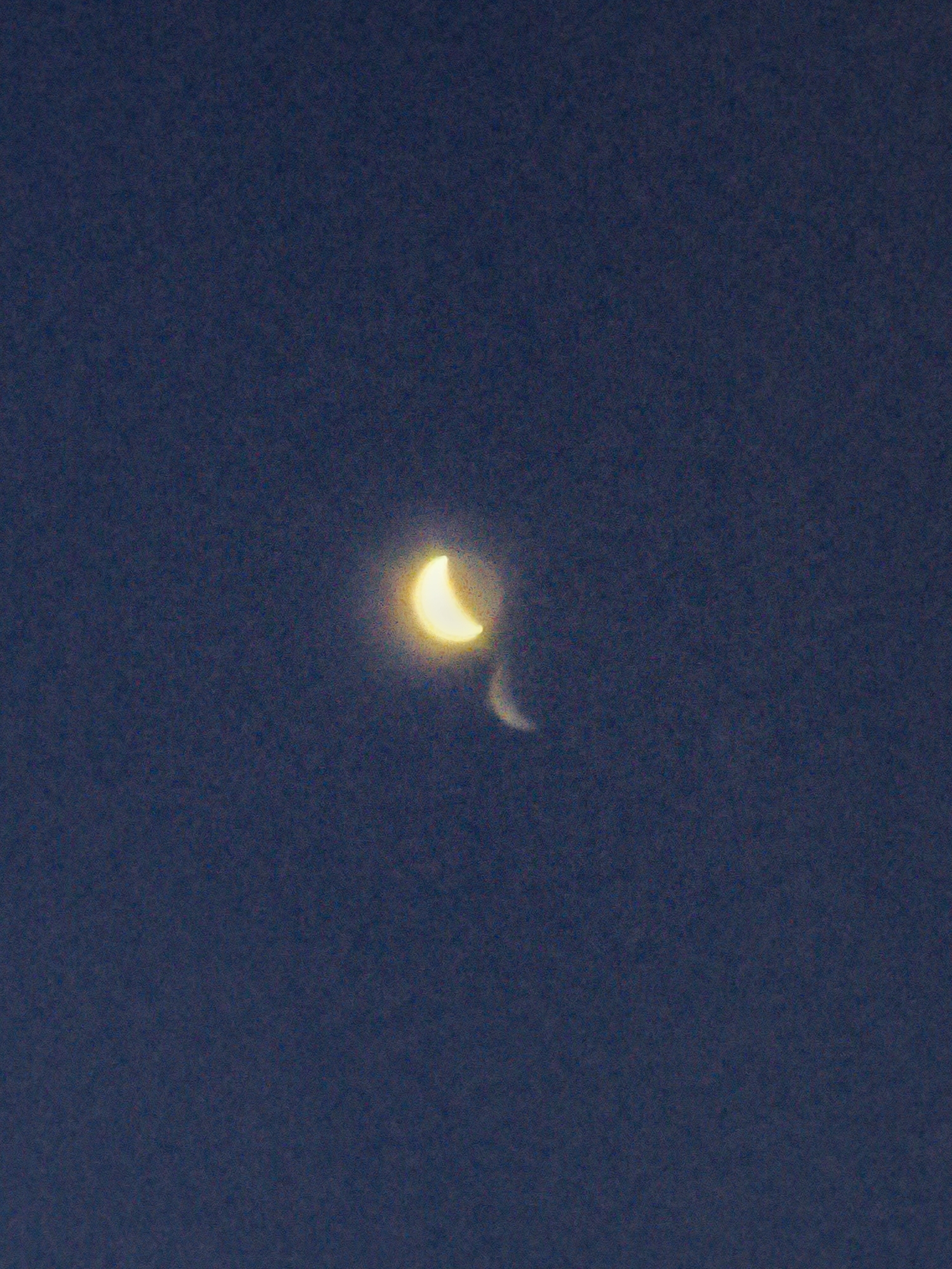 The image shows a sliver of the crescent moon in a dark blue night sky. There is a faint, lighter crescent shape to the right of the main crescent, possibly an artifact or a very faint Earthshine. The image appears to be taken from the Lachish Regional Council, Israel, based on the provided context. The sky is clear and dark, indicating a nighttime scene. There are no people or man-made structures visible.