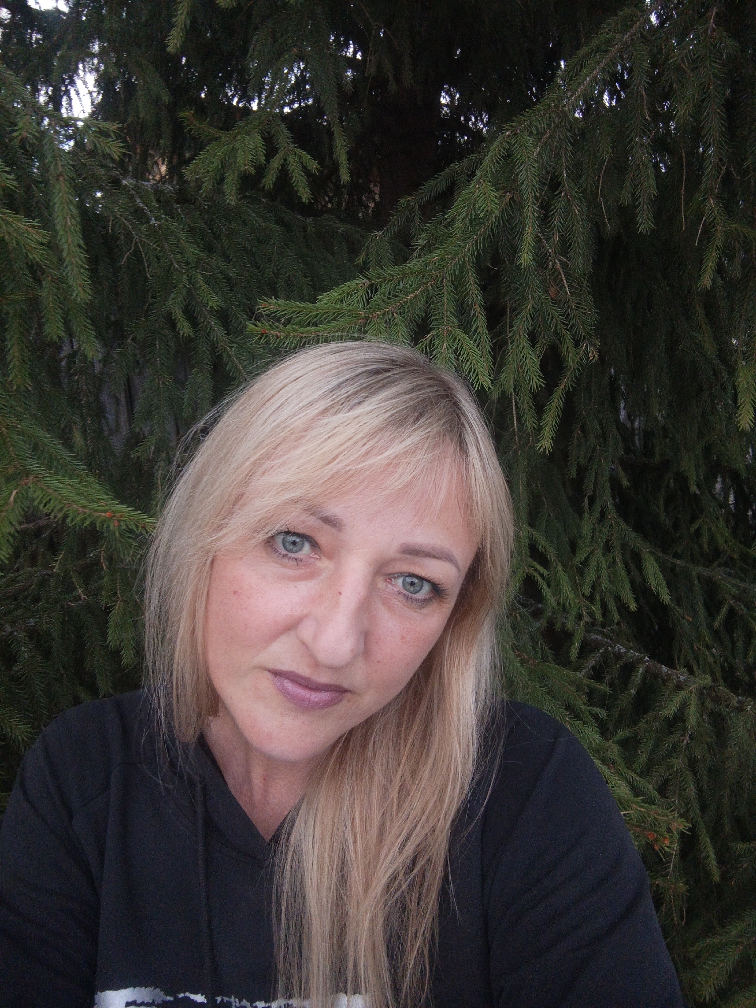 A woman with blonde hair and blue eyes stands in front of an evergreen tree. She is wearing a black hooded sweatshirt. The scene is located in the city of Chaikovsky, Russia.