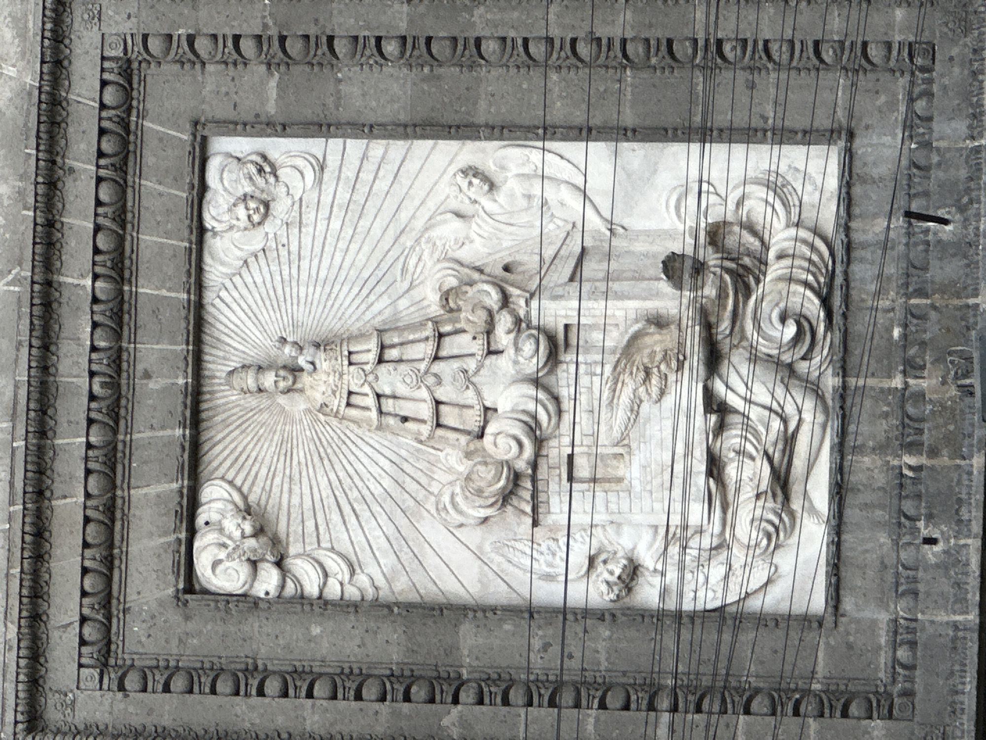 The image shows a rectangular, carved bas-relief panel set into a gray stone structure, possibly a ceiling or wall. The carving depicts religious iconography, featuring a multi-tiered structure resembling a tower or shrine emanating rays of light. Above the tower is a figure of an angel. A second angel is present. The bas-relief is surrounded by an elaborate, multi-layered frame with repeating patterns. Electrical wires cross the panel. The style of the artwork suggests a religious building. The architecture and imagery suggest this is located in Mexico City, Mexico.