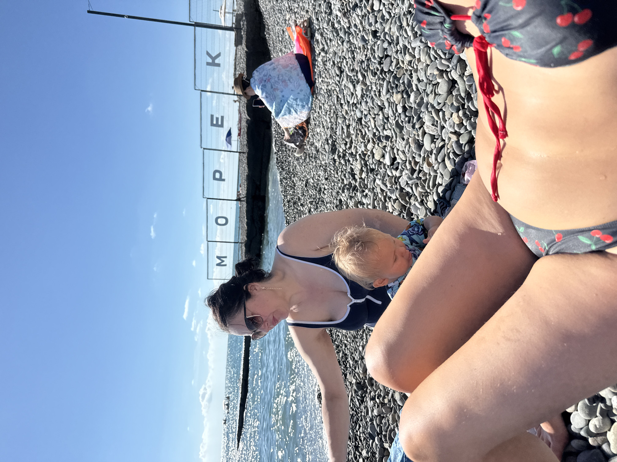 The image depicts a pebble beach in Adlerskiy, Russia, on a sunny day. In the foreground, a person's legs are visible, wearing a bikini with a cherry pattern. To the left, a woman in a dark blue swimsuit and sunglasses is interacting with a baby. The baby is wearing a patterned outfit. In the background, a person wearing a hat and a light blue wrap is sitting on an orange mat, looking at their phone. There is a fence with large letters spelling out "M O P E K" in the background, possibly indicating a location name. The sea is visible with gentle waves and sunlight reflecting on the water. Additional beach umbrellas and a pier can be seen in the distance. The beach is covered with grey and dark pebbles.