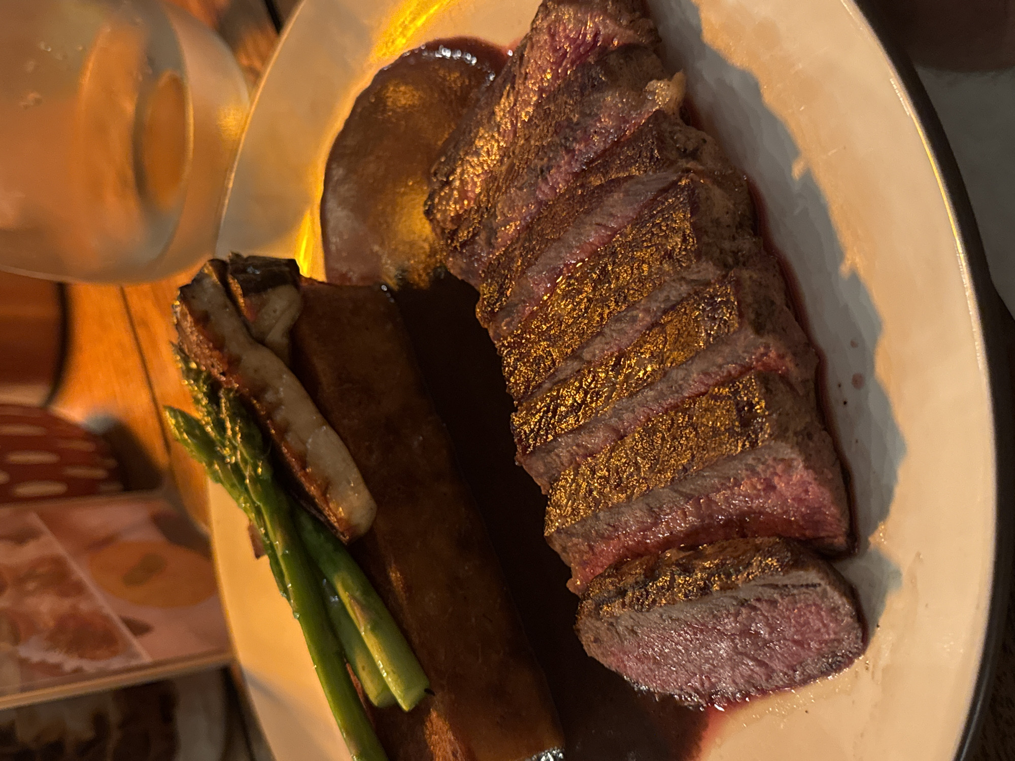 A plate of sliced steak with asparagus, a potato cake, and sauce is presented on a white plate. The steak is rare and appears to be seared on the outside. The asparagus is green and positioned near the potato cake. The sauce is a dark brown color. A glass of clear liquid sits next to the plate. The image was taken indoors in the city of Kuta Selatan, Indonesia.