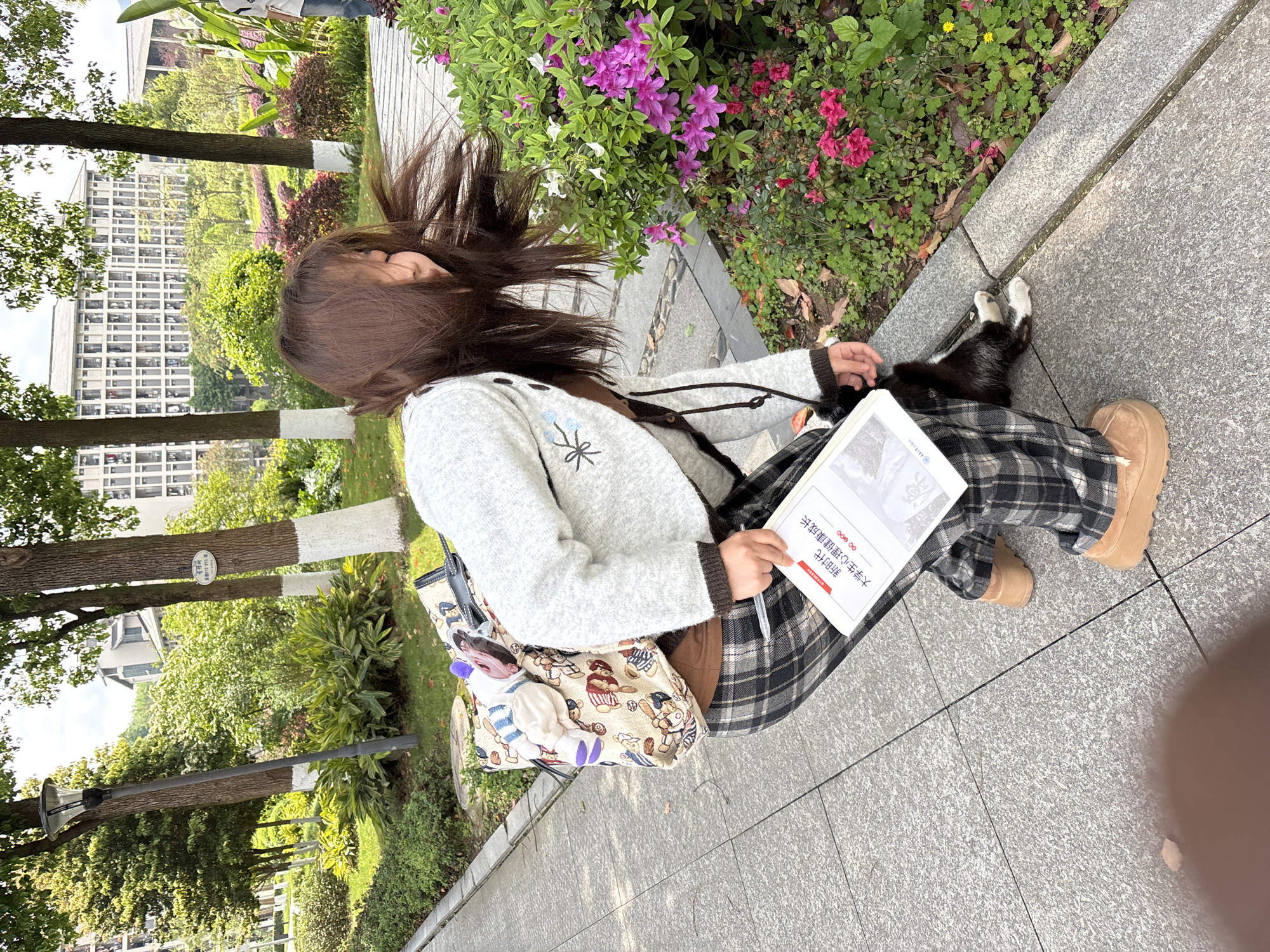 A young woman with dark hair sits on a concrete pathway alongside a flowerbed, holding a book titled "大学生心理健康成长" (roughly translated as "University Students' Mental Health Growth") and a pen. She wears a light grey cardigan, a plaid skirt or pants, and brown boots. A patterned bag rests at her side. The location appears to be in Jiangjin, China, based on the Chinese characters on the book. Trees and a building are visible in the background. The ground is paved with grey stone. A cat with white paws is sitting near her right leg.