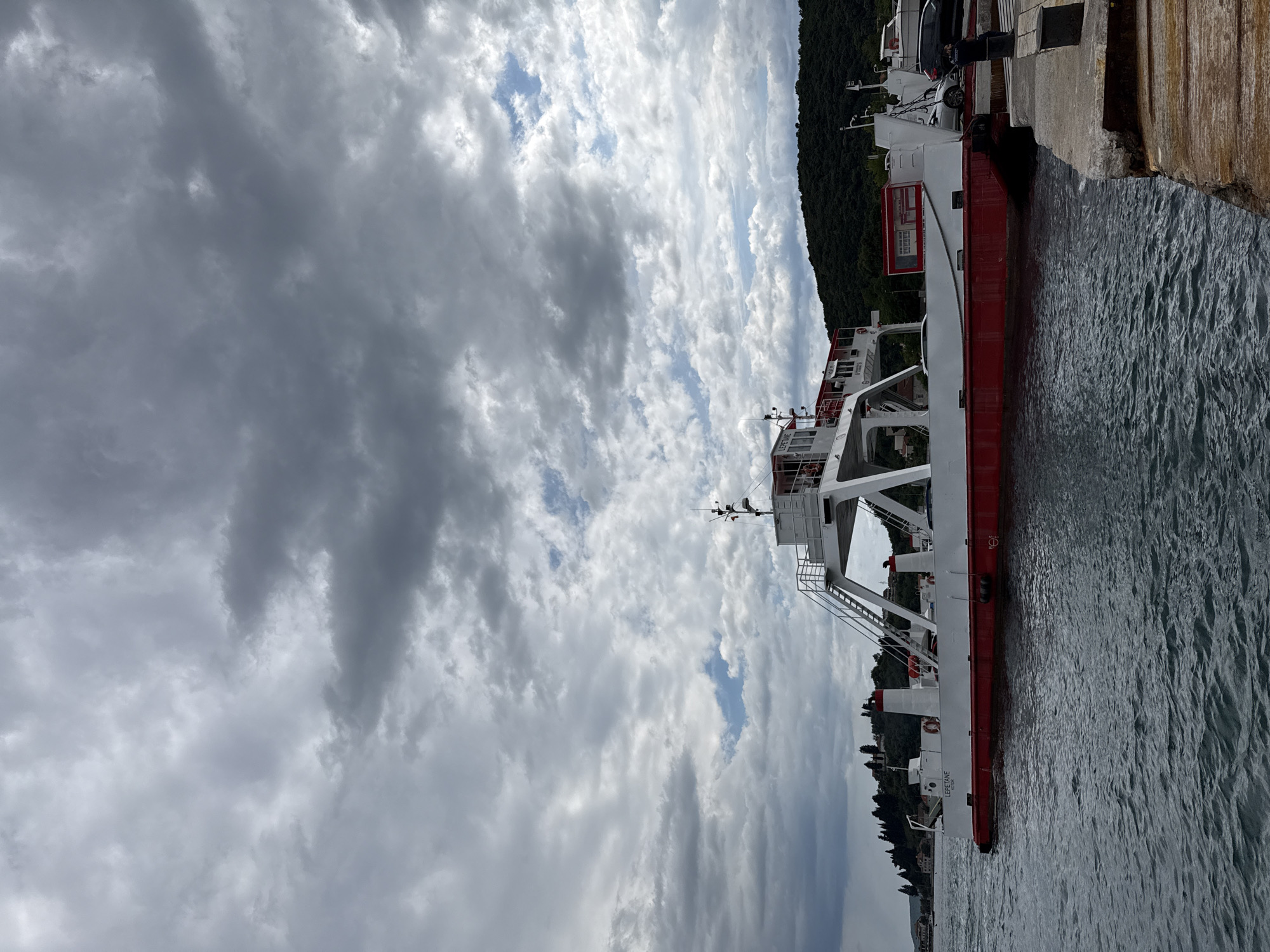 The image depicts a ferry docked in Đurići, Montenegro. The ferry is white with a red stripe along its hull and several levels. It is moored to a concrete pier. The water surrounding the ferry is dark, suggesting overcast weather. The sky is filled with gray and white clouds, partially obscuring the sun. Lush green trees and vegetation line the background, indicating a coastal environment. The ferry is labeled "LEPETANE."