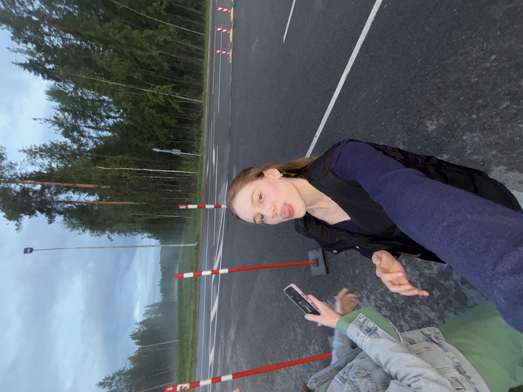 The image depicts two individuals near a road, likely in Arfa, Russia. The foreground features a young woman with light brown hair, wearing a dark vest over a navy top, and a dark blue knitted sleeve. She is sticking her tongue out playfully at the camera. To her left, another person, mostly obscured, wears a light blue denim jacket over a green garment. This person holds a pink smartphone. Red and white striped traffic cones are visible on the asphalt road and its shoulder. A forest lines the background under a cloudy sky. The overall scene suggests a casual, outdoor moment.