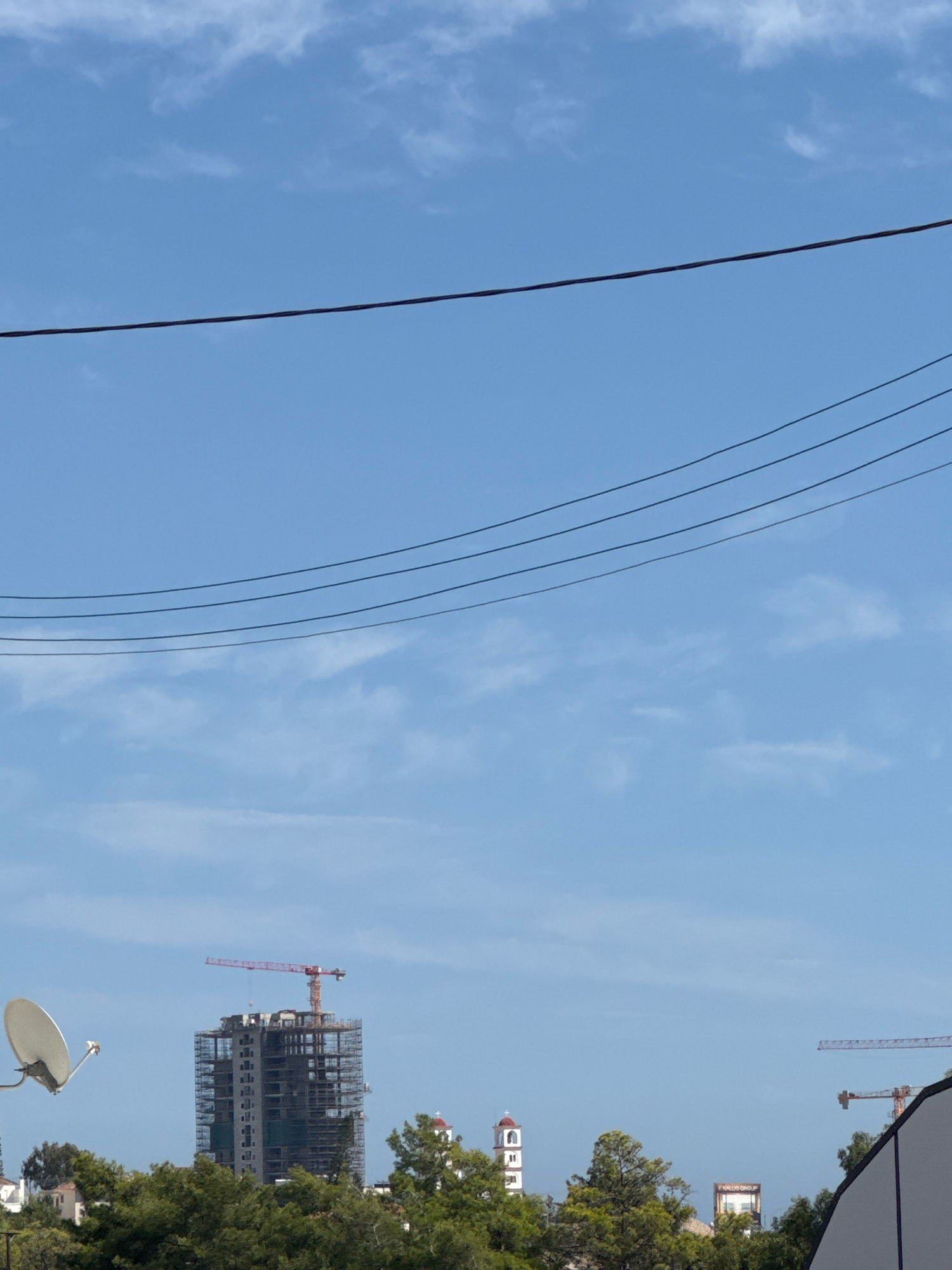 The image shows a bright, clear sky with scattered clouds over a cityscape. In the foreground, power lines are strung across the frame. In the middle ground, a tall, unfinished building is under construction, surrounded by scaffolding and topped with a red construction crane. To the left of the building, a satellite dish is visible. Further in the distance, a tower with a red dome and cross, resembling a church or bell tower, stands among trees. Another construction crane is visible on the right side of the image. The scene appears to be from an urban environment, likely Germasogeia Municipality, Cyprus, given the context. The lighting suggests it is daytime.