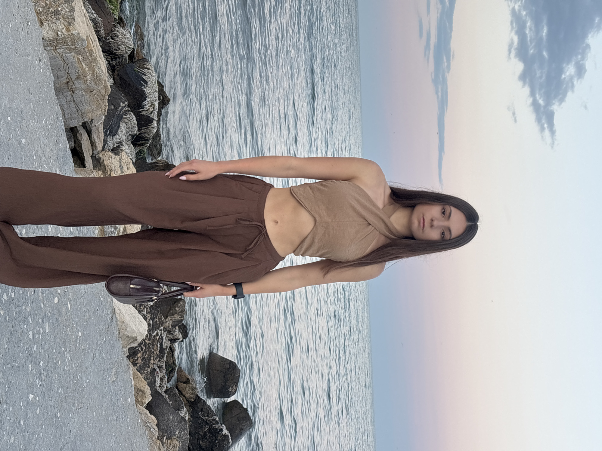 A young woman is standing on a rocky shore next to the sea. She is wearing a brown top and brown pants. The sea is calm, and the sky is a mix of blue and pink, suggesting it is either sunrise or sunset. The woman is holding a dark-colored handbag. The rocks in the foreground are varied in color and texture, with some being light gray and others dark. The water appears to be a pale blue. The scene suggests a peaceful and serene moment by the sea. The woman has long, dark hair and is looking towards the camera. The overall mood of the image is tranquil and natural.