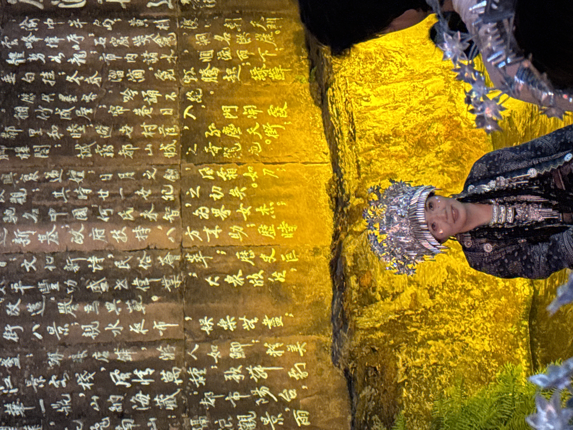 A medium shot captures a section of an ancient stone wall inscribed with Chinese characters, illuminated by a warm yellow light. The characters, rendered in white, are densely packed and appear to be in an aged calligraphic style. In the foreground, a person wearing a traditional ornate silver headdress and intricate dark clothing smiles towards the camera. The headdress features numerous hanging silver ornaments. To the right of the person, another individual is partially visible, also adorned with silver accessories. The background wall is rough and textured, suggesting it's a natural rock face or an ancient structure. The scene appears to be outdoors, possibly in a historical or cultural site within Yuzhong District, China, given the presence of ancient Chinese inscriptions and traditional attire.